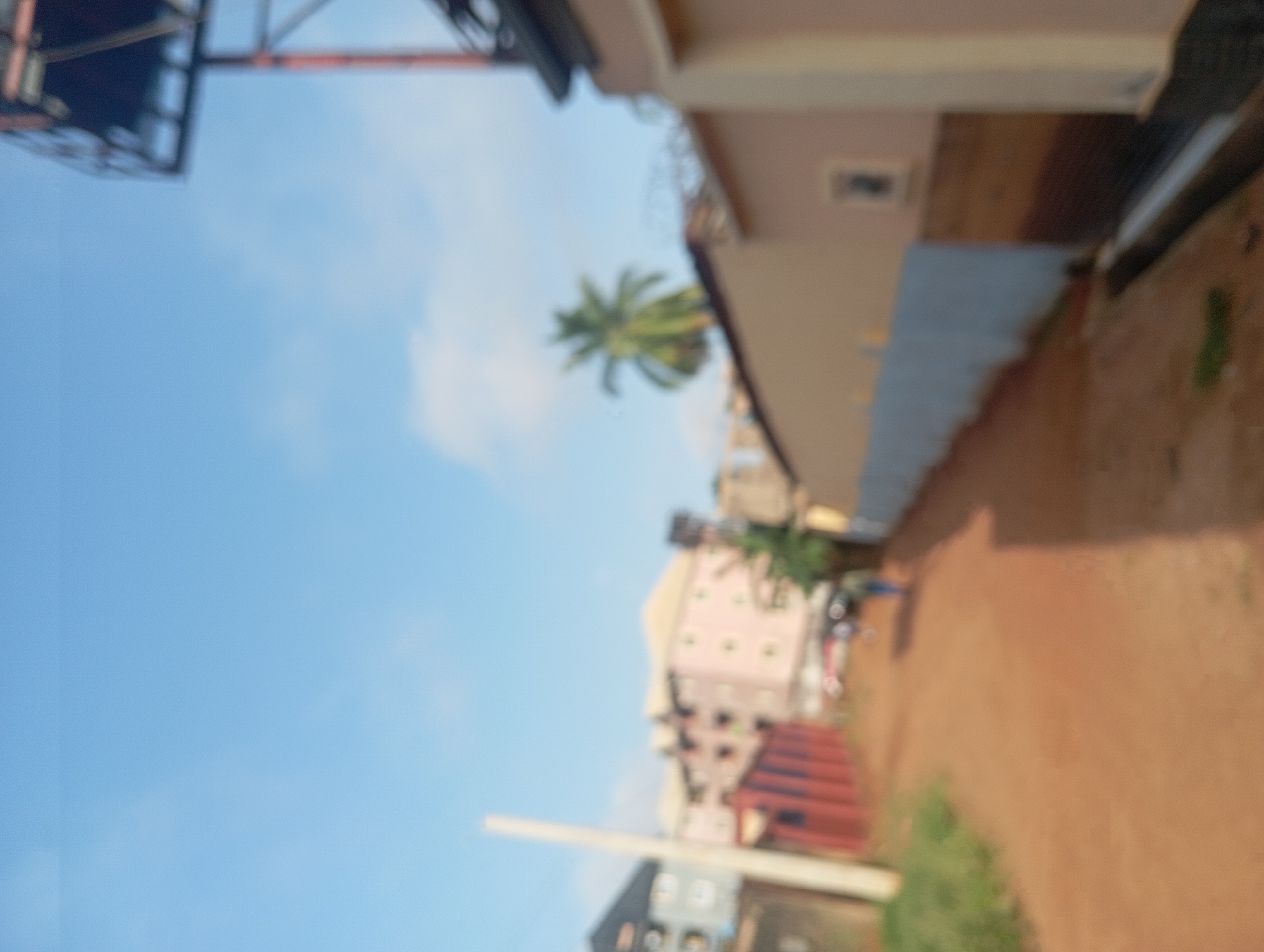 The image shows an outdoor scene in Nkpor, Nigeria. A blurry red dirt road is visible on the right side of the image. Buildings of various heights and colors are visible in the background. A palm tree is visible near one of the buildings. The sky is blue with some clouds. A metal structure is visible at the top of the image. The image is generally out of focus.