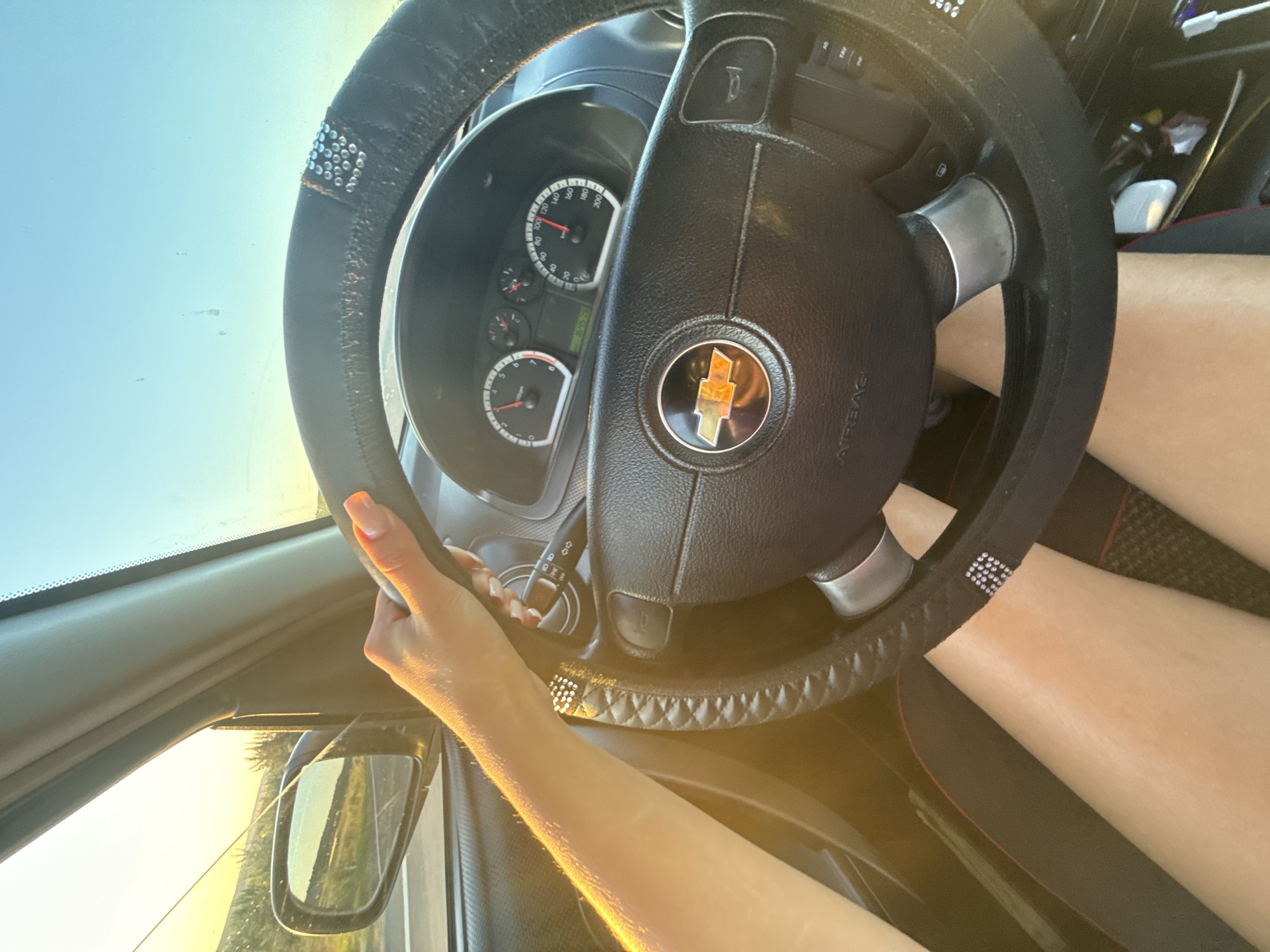 An interior shot captures a person's hand, with bright orange nail polish, gripping the steering wheel of a car. The steering wheel is black with decorative rhinestones and a Chevrolet logo in the center. The dashboard displays the speedometer, indicating the car is traveling at approximately 90 km/h. The sun's golden light streams in through the windshield and side window, casting a warm glow on the scene and illuminating the person's legs, which are visible below the steering wheel. Outside the car, a rural landscape with trees and a distant road is visible in the side mirror, bathed in the soft light of either sunrise or sunset. The overall impression is one of a leisurely drive through the countryside, possibly on the outskirts of Cimișlia, Moldova.