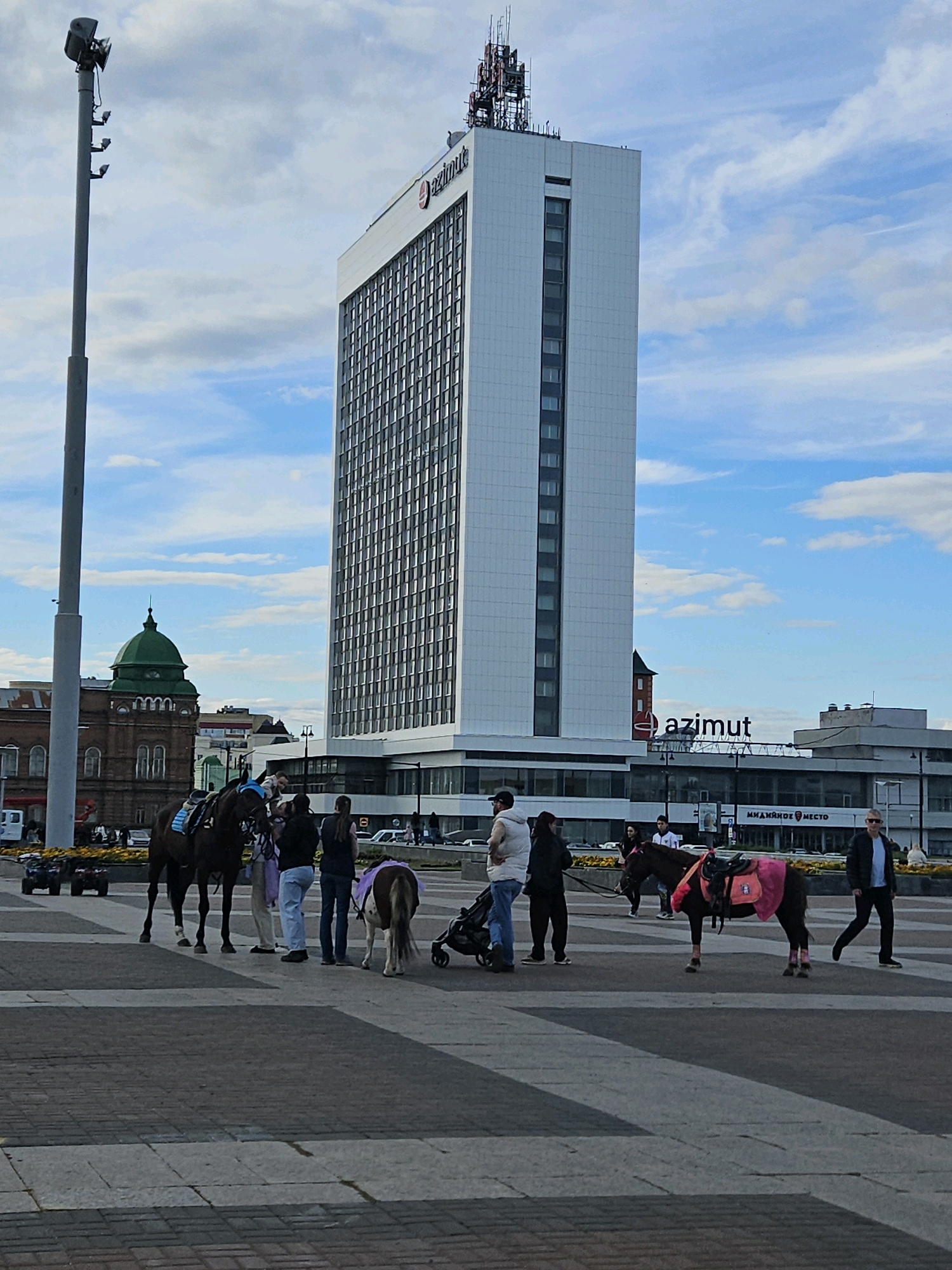 In Ulyanovsk, Russia, a group of people stand in a paved square in front of a tall, modern white building with the "azimut" logo prominently displayed. To the left of the modern building is a smaller, older brick building with a green dome. In the foreground, two horses are present: a larger dark brown horse and a smaller pony. The pony is adorned with a pink blanket and leg wraps. Several individuals are interacting with the horses, and a stroller is visible. In the background, there are more buildings and vehicles, indicating an urban setting. The sky is partly cloudy.