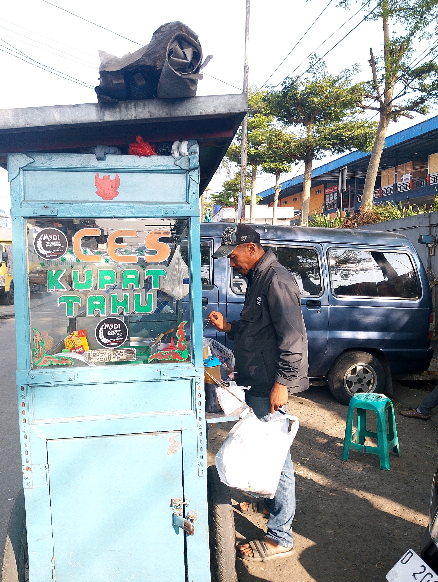 A street vendor in Lembang, Indonesia, stands beside his mobile food cart. The cart is light blue and displays the words "CES KUPAT TAHU" in large, colorful letters. The vendor, wearing a black jacket and a baseball cap, is in the process of serving a customer or preparing an order. He is holding a white plastic bag. A dark blue van is parked behind the cart. The environment appears to be outdoors, with trees and buildings visible in the background, suggesting a street or market setting. The ground is paved, and there is a green plastic stool nearby. The lighting indicates it is daytime, possibly late afternoon given the shadows.