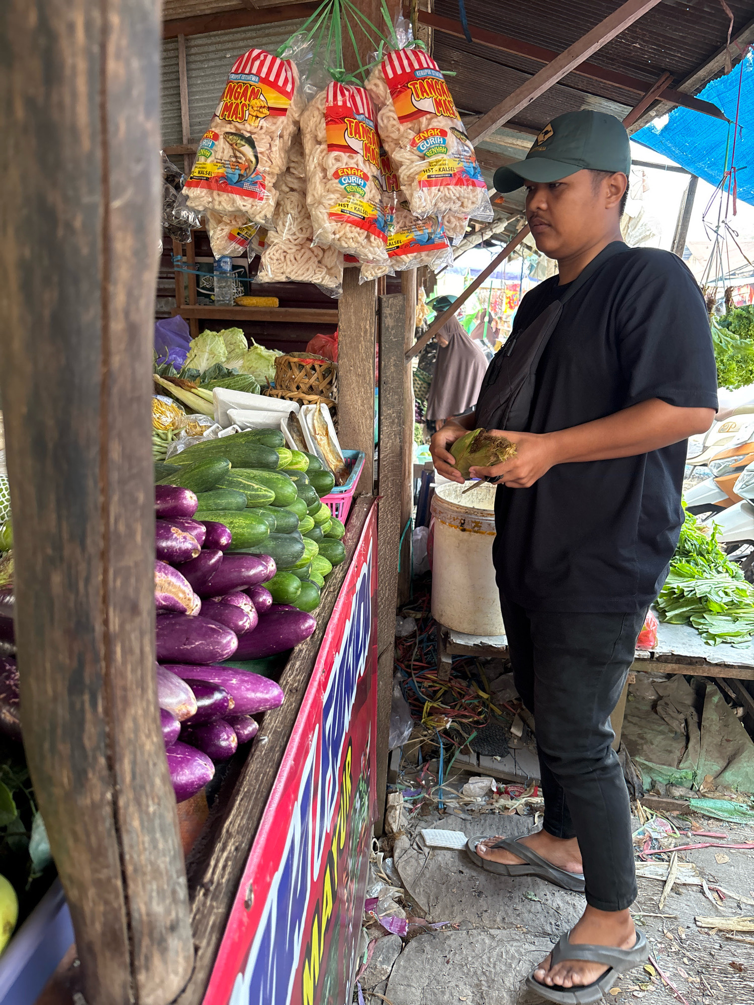 In Banjarmasin, Indonesia, a person wearing a green cap, black t-shirt, and black pants stands at a market stall. The stall displays an assortment of fresh vegetables, including eggplants and cucumbers. Hanging from the stall are several bags of snacks. The person is holding a green object, possibly corn, in their hands. The market environment is visible with various items and debris on the ground, indicating a bustling outdoor market scene.