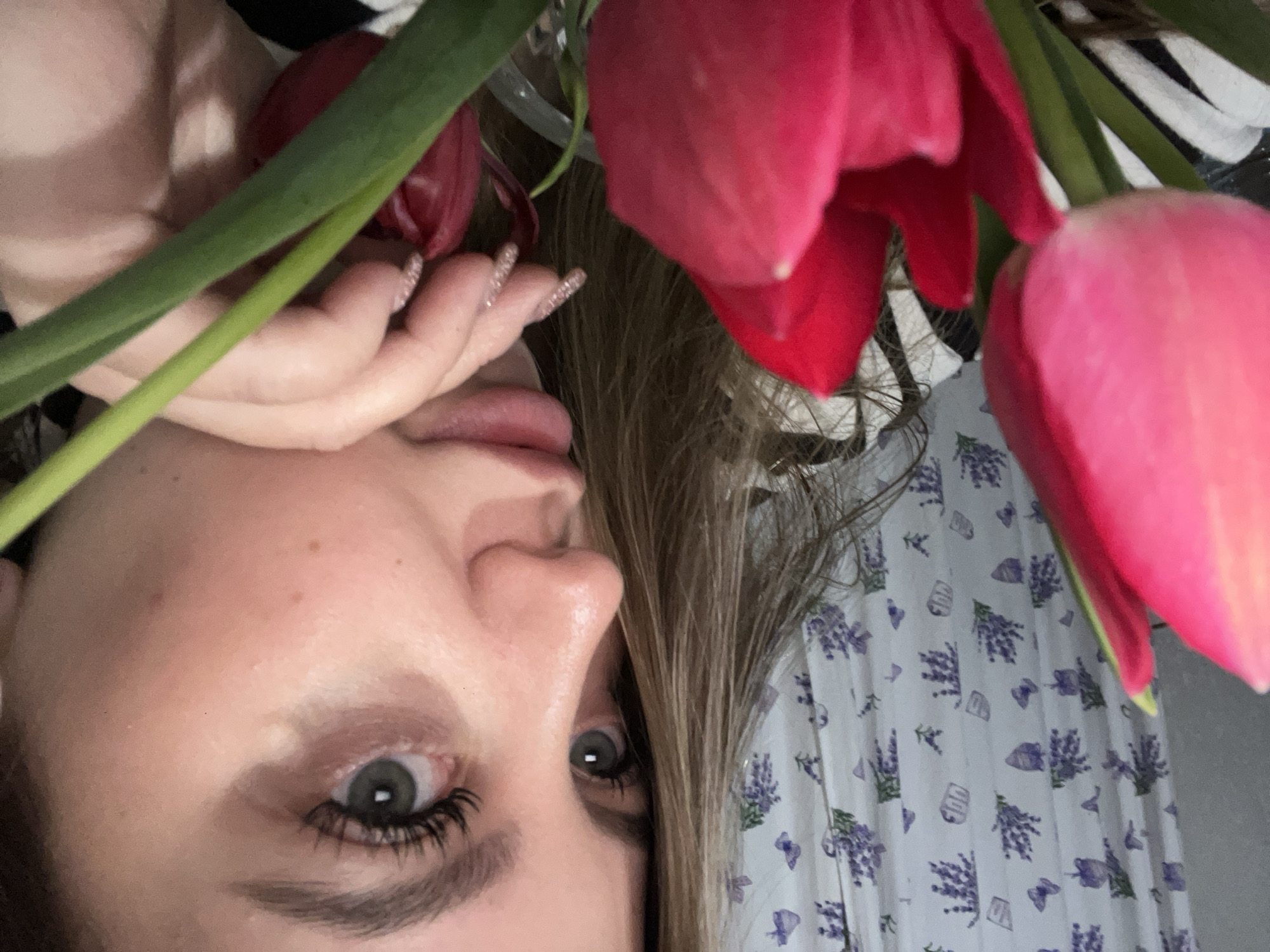 A selfie shows a woman's face next to red and pink tulips with green stems. The woman has light skin, light brown hair, green eyes, and long eyelashes. Her hand is near her face, and her nails are decorated with glitter. The background includes a white curtain with a lavender floral pattern and a black and white striped fabric. The location is not discernible from the image and the reference to the city of Novopokrovskaya, Russia, cannot be confirmed.
