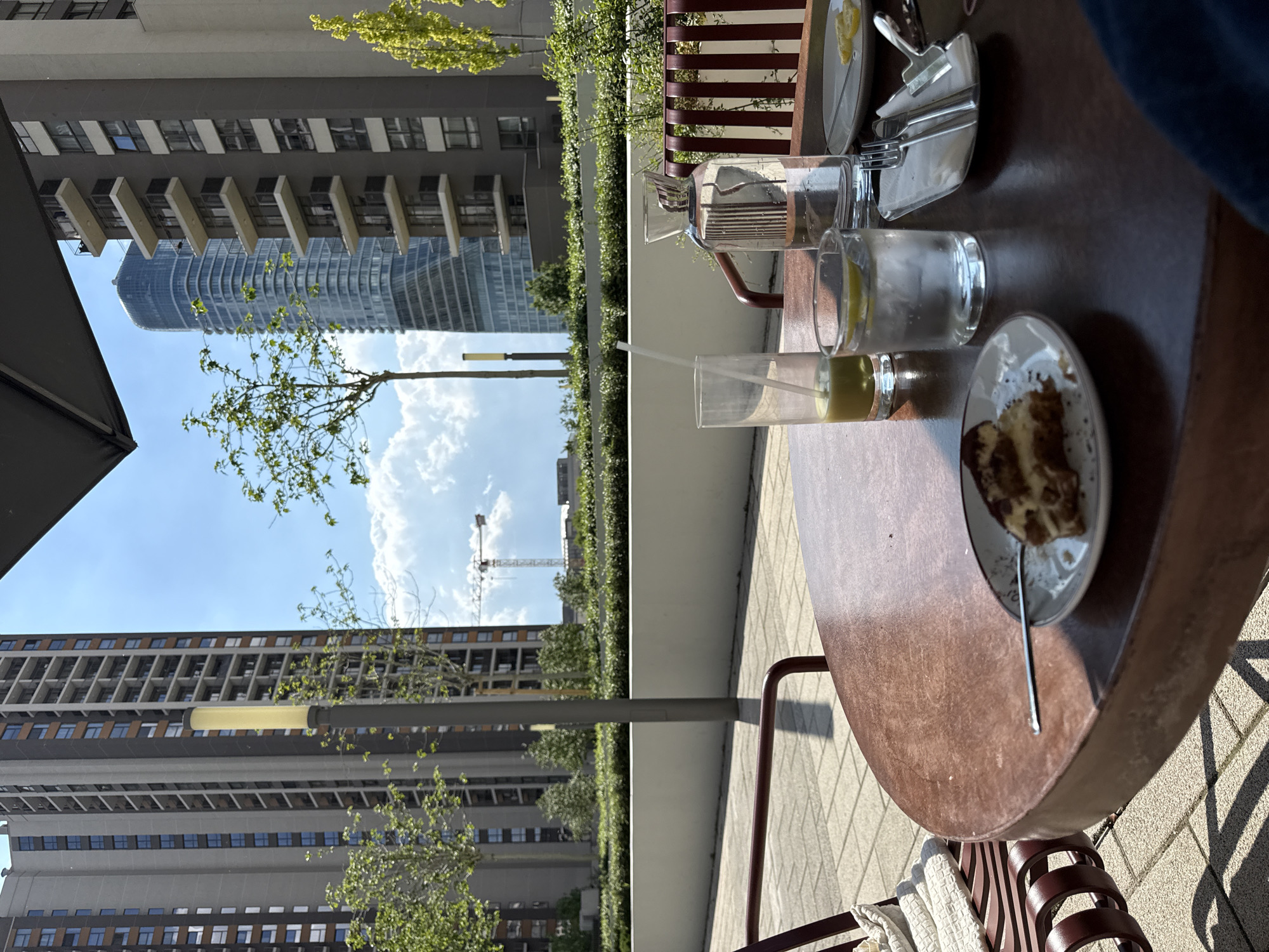 Here is a description of the image:
The image shows a table setting at an outdoor cafe or patio with buildings visible in the background. On the table, there are plates, cutlery, glasses with drinks, and a partly eaten cake. Beyond the table and patio area, there is a view of the city, including modern buildings and a construction crane under a blue sky with some clouds.
Based on the architecture of the buildings in the background, it's plausible that the location is in Belgrade, Serbia, specifically around the Belgrade Waterfront area.