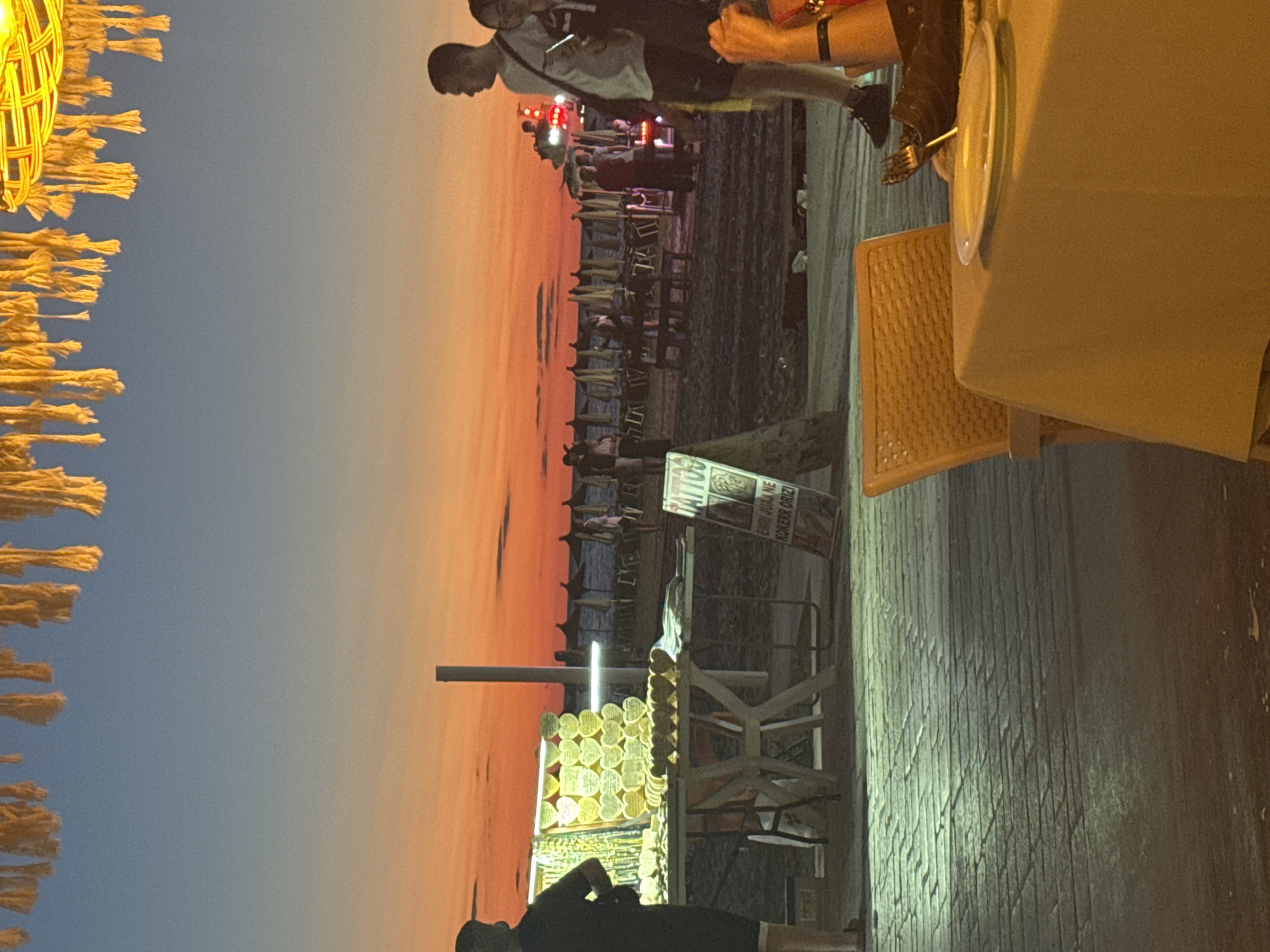 The image captures a scene at dusk on a beach in Golem, Albania. In the foreground, a table is set with a yellow tablecloth and a plate, suggesting an outdoor dining area. To the left, a vendor's stall displays numerous heart-shaped wooden items, likely souvenirs, illuminated by bright lights. A person stands behind the stall, facing away from the camera. In the background, the beach is visible with rows of closed umbrellas and lounge chairs, indicating the end of the day. Several people are scattered across the sand, some walking and others gathered near what appears to be a food stand or a kiosk. The sky displays a vibrant gradient of orange and blue, characteristic of a sunset. The overall atmosphere is relaxed and suggestive of a tourist destination.