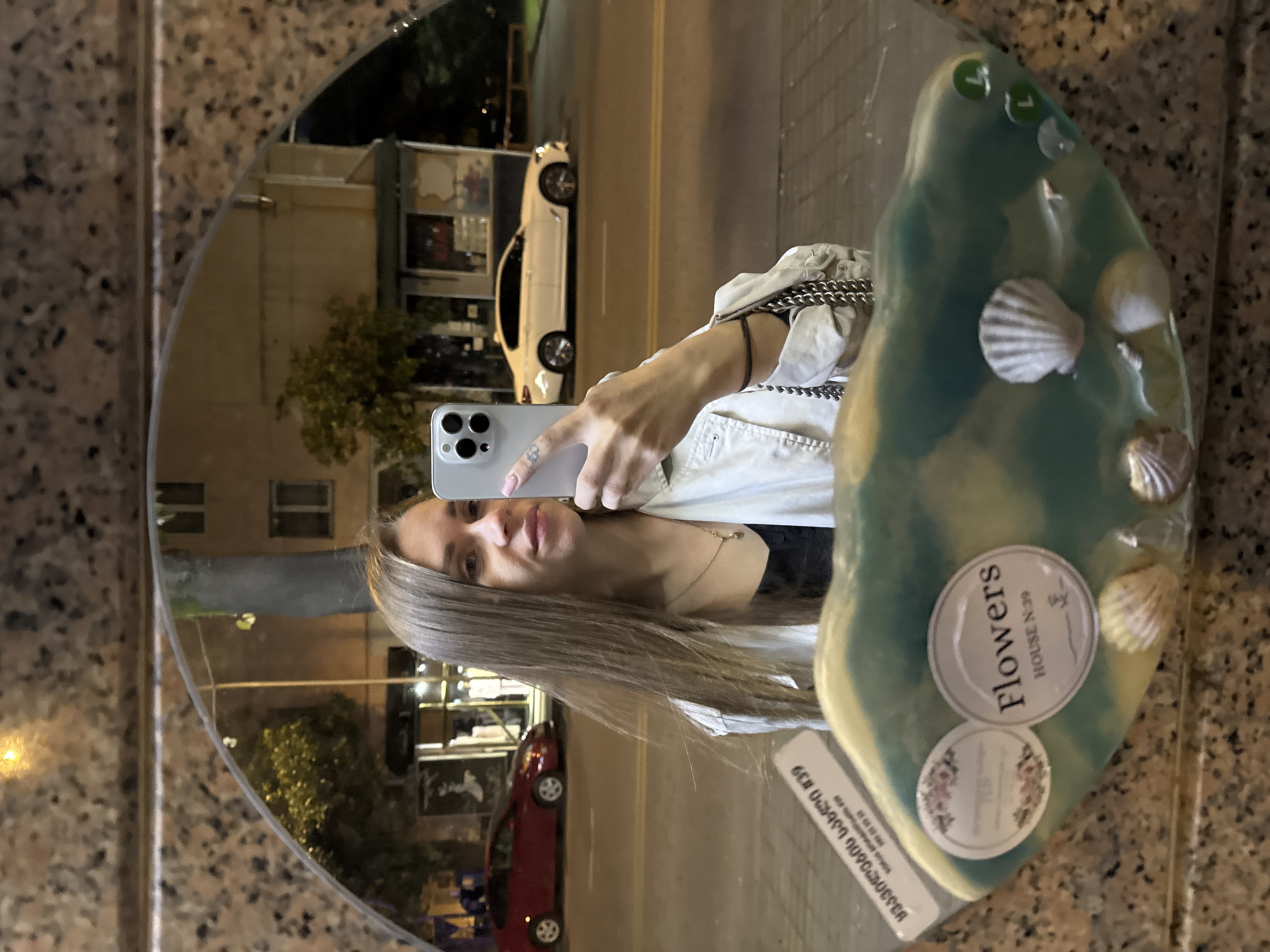 A young woman is captured in a nighttime mirror selfie, reflecting a bustling street scene in Batumi, Georgia. She holds a light-colored smartphone with a triple camera array to take the picture, her face and upper body visible. She has long, straight, light brown hair and wears a light-colored jacket over a dark top, accessorized with a delicate necklace and a bracelet.
The circular mirror, possibly framed with a speckled granite or marble border, reflects a street illuminated by various lights. In the reflection, a white sedan and a red sedan are parked or driving along the road. Buildings and some trees line the street, suggesting an urban environment at night.
A distinctive decorative element is affixed to the right side of the mirror. This object features a swirled blue-green and yellow-white pattern, resembling resin or glass, and has small seashells embedded within it. Two circular stickers are visible on this decoration. The top sticker reads "Flowers" and "HOUSE N.39", with smaller, unreadable text below. The bottom sticker is partially obscured but appears to be another floral-themed label. Below these, a rectangular sticker with Georgian script is visible, starting with "639" and including the word "სუვენირების" (souvenirs), followed by what appears to be a phone number. The presence of Georgian text and the reference to souvenirs further reinforces the location context of Batumi.
The woman exhibits a neutral to slightly smiling expression as she photographs herself. The overall scene conveys a casual moment captured in a reflective surface, blending personal interaction with an external urban environment.