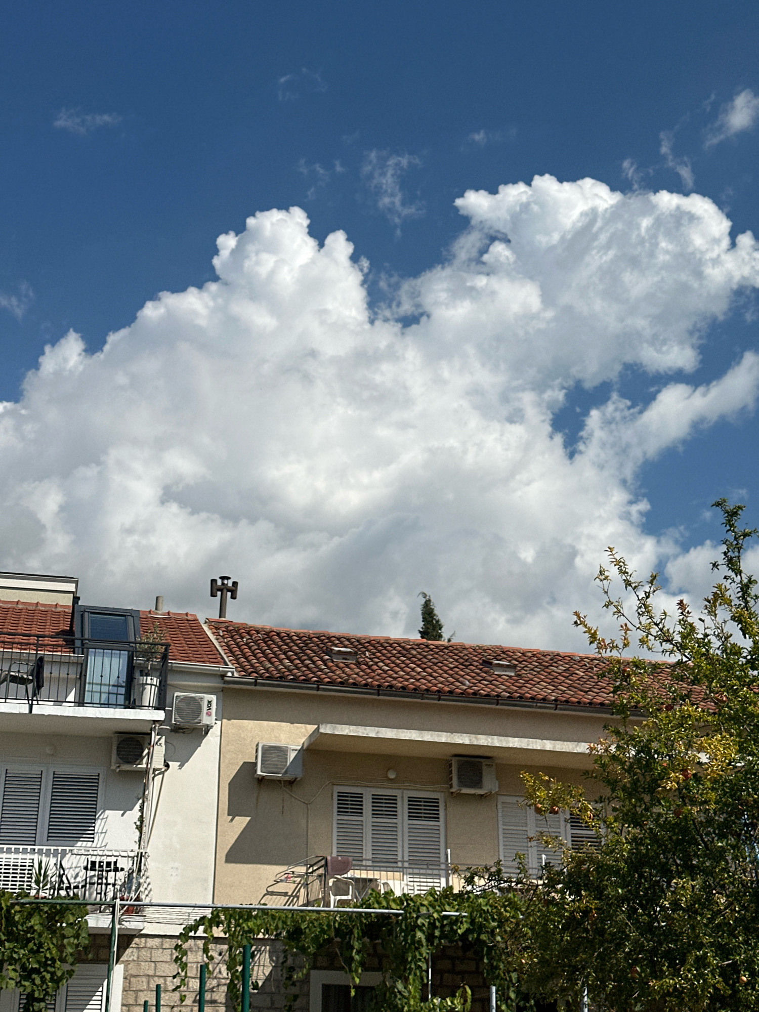 The image captures a sunlit scene in Budva, Montenegro, with a prominent sky filled with large, fluffy white clouds against a deep blue backdrop. In the foreground, the terracotta tiled roofs of buildings are visible, casting shadows. On one of the roofs, a tall, dark structure with multiple horizontal elements resembling a stylized antenna or siren stands out. Adjacent to this roof, a building with white shutters and multiple air conditioning units is visible. Lush green foliage, including vines and a tree with some red fruit, partially obscures the lower part of the buildings. The overall impression is of a bright, possibly warm day in a residential area.