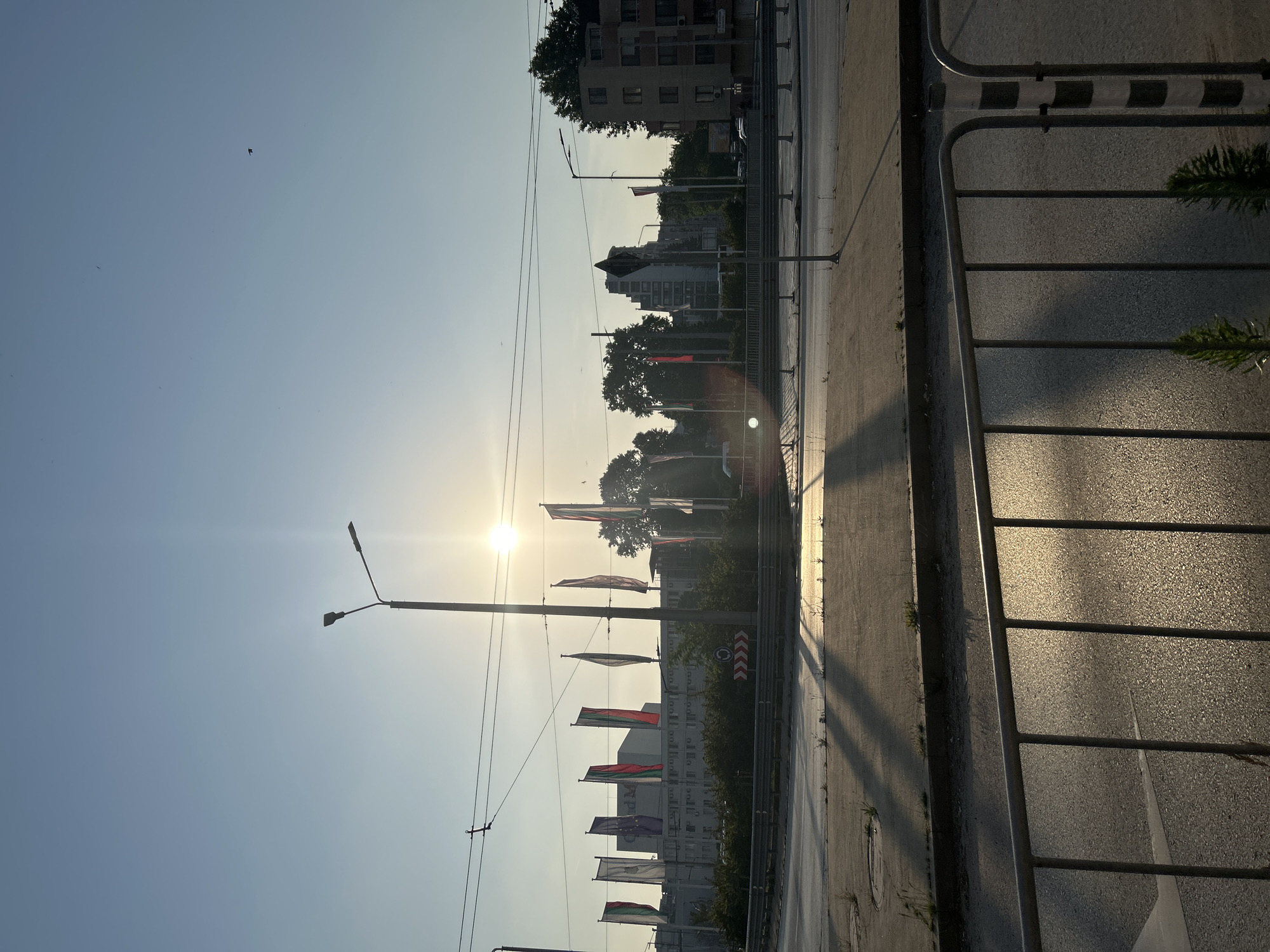 This image depicts an urban landscape in Varna, Bulgaria, at a time when the sun is low in the sky, either early morning or late afternoon, as evidenced by the long shadows and direct sunlight. The sky is clear and light blue, with a prominent sun in the upper mid-right creating a bright lens flare. Several small dark specks, likely birds, are visible against the sky.
In the immediate foreground, a metal barrier fence runs horizontally across the bottom of the frame. Beyond the fence, a paved road surface reflects the sunlight, and distinct shadows are cast from the fence and other objects towards the lower right. Small green plants grow sparsely on the pavement near the fence. A black and white striped pole is visible on the right edge.
The midground features a multi-lane roadway or intersection, appearing largely empty of traffic. A prominent street light with two lamps stands near the center-left. Overhead power and trolley lines crisscross the scene. A central island or median is covered with low green bushes, partially obscuring a circular road sign and a red and white chevron sign indicating a directional change or hazard.
In the background, on the left side of the frame, stands a large, light-colored building with the letters "Grand M" visible on its upper facade. In front of this building, several tall, rectangular flags displaying the vertical stripes of red, white, and green, characteristic of the Bulgarian national flag, are visible. To the right, further buildings, including a multi-story structure with balconies and another streetlight, are partially visible through trees. The overall scene appears to be an urban intersection or roundabout. No people are discernible within the frame.