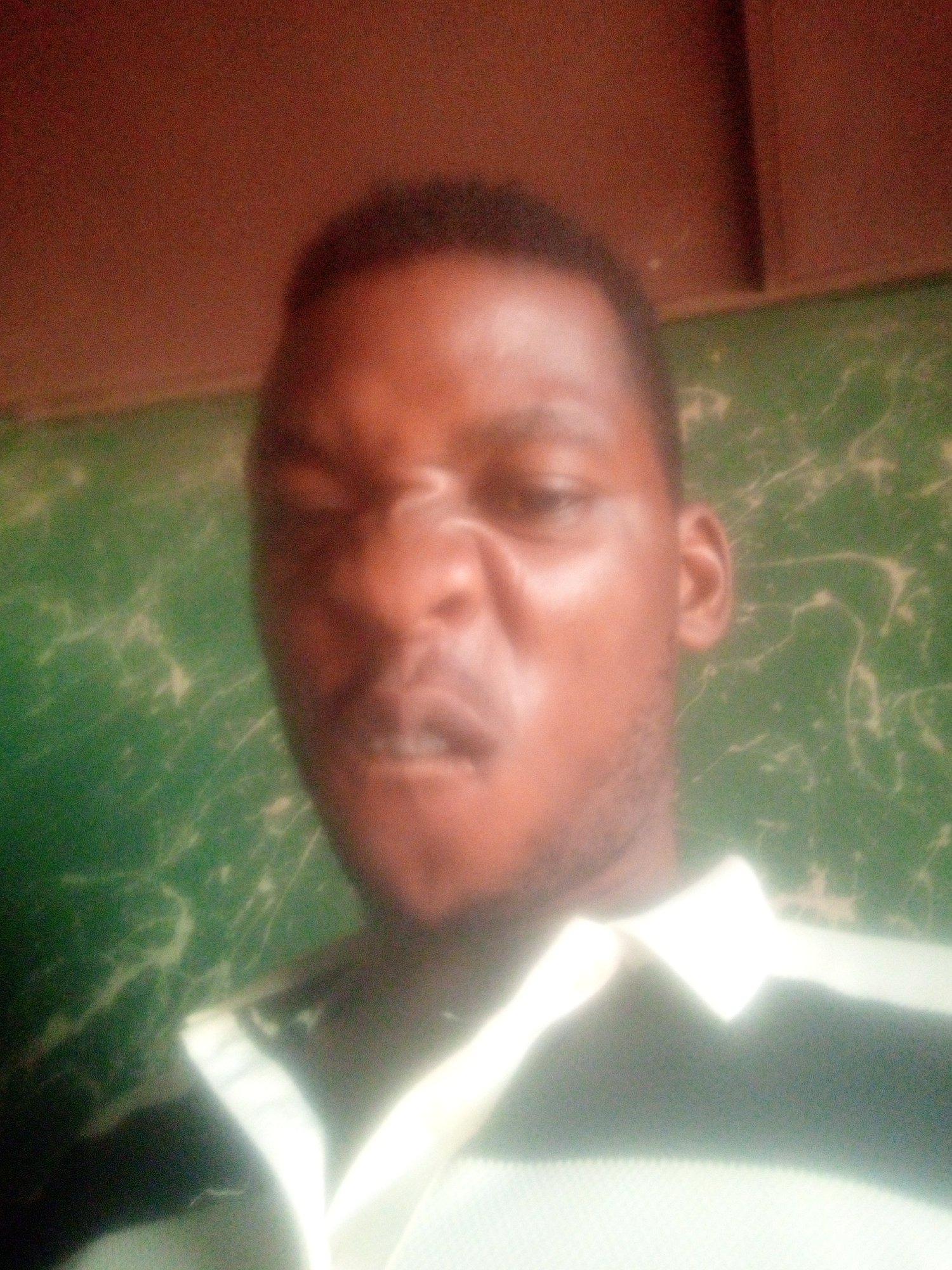 The image features a close-up, slightly blurry portrait of a person, likely a man, with dark skin and short dark hair. He is positioned in the center of the frame, looking directly at the camera with an exaggerated, humorous expression. His eyes are narrowed, his nose is scrunched up, and his mouth is slightly open, revealing some teeth. A faint amount of facial hair is visible around his jawline.
He is wearing a collared shirt, which appears to be a polo shirt, with a white collar and a body that has black and white horizontal stripes, though the stripes are largely out of focus due to the shallow depth of field and blur.
The background is divided into two main sections. The upper part is a solid, dark reddish-brown color, possibly a ceiling or an upper wall section. The lower and larger portion of the background is a vibrant green wall with lighter, white or light green, mottled patterns, resembling a painted or textured surface.
The lighting seems somewhat soft and diffused, contributing to the overall slightly grainy and out-of-focus quality of the photograph. No specific location cues beyond an indoor setting are discernible, and there is no visible text. The image conveys a playful or lighthearted mood due to the subject's expression.