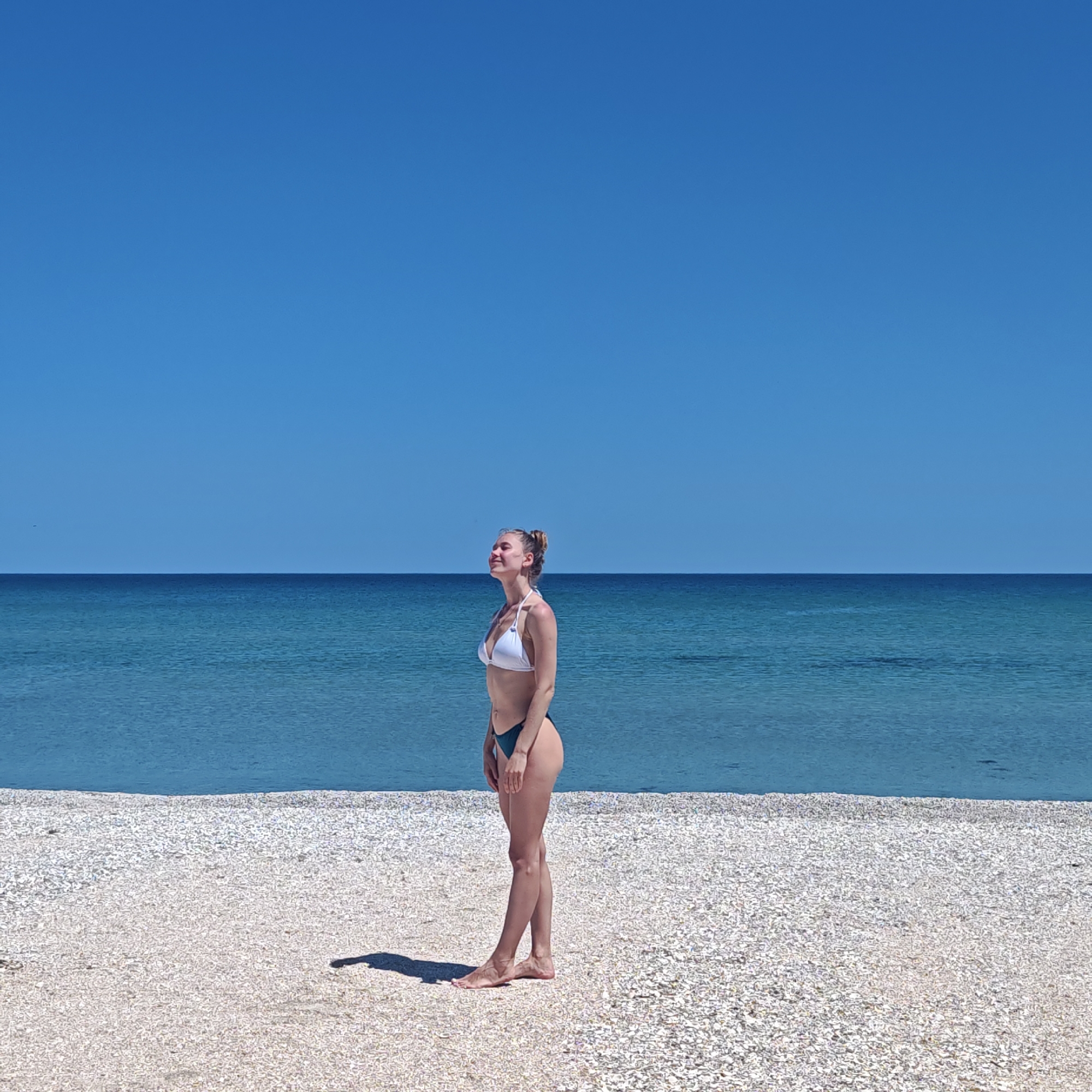 A woman stands on a beach composed of small white shells under a clear blue sky. The horizon line divides the sky and the blue sea in the background. The woman is facing to the right and is wearing a white bikini top and dark blue bottoms. She has her eyes closed and head tilted up as if sunbathing. Her shadow is cast on the beach to the left. The location is Unknown, Unknown.