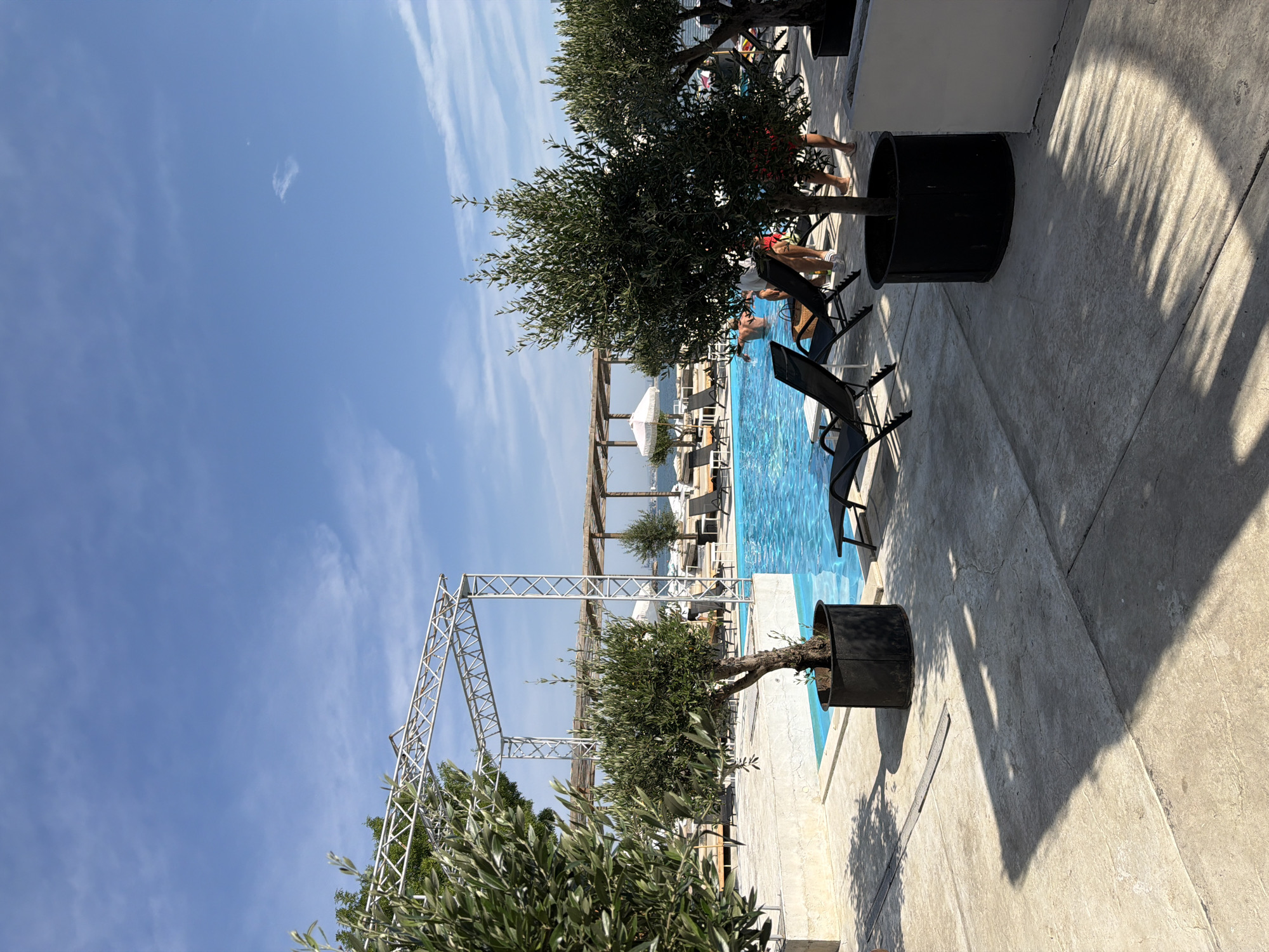 The image depicts an outdoor scene at a resort or hotel with a swimming pool in Burgas, Bulgaria. The swimming pool is filled with bright blue water and is partially visible on the right side of the frame. Several lounge chairs are positioned around the pool area, with some occupied by people. Two olive trees in large black pots are prominent in the foreground, one on the left and another in the center, casting shadows on the concrete ground. A metal structure, possibly for staging or shade, is visible behind the central olive tree. In the background, there are additional lounge chairs, umbrellas, and a view of what appears to be the sea or a large body of water. The sky is clear and blue with a few wispy clouds. People are seen in and around the pool, some swimming and others relaxing. The overall atmosphere is one of leisure and summer vacation.