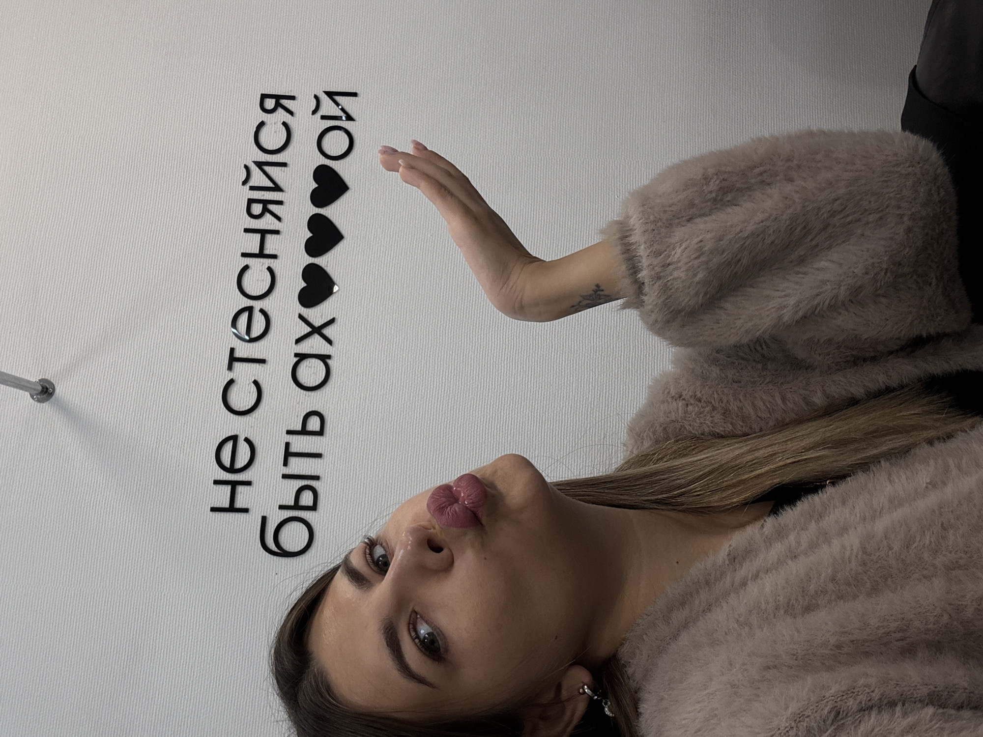 The image shows a young woman with long, blonde hair, wearing a fluffy, beige cardigan over a dark top. She is making a kissing face and looking towards the camera. Her right hand is raised with fingers slightly curled. A tattoo is visible on her forearm. The background is a textured white wall with black lettering that reads "не стесняйся быть ах♥ ♥ ♥ ой". This translates to "Don't be shy to be hot." A metal fixture is visible at the top of the wall. The image appears to be taken indoors, likely in a residential or commercial space. There is no indication of the specific location within the city of Chaikovsky, Russia.