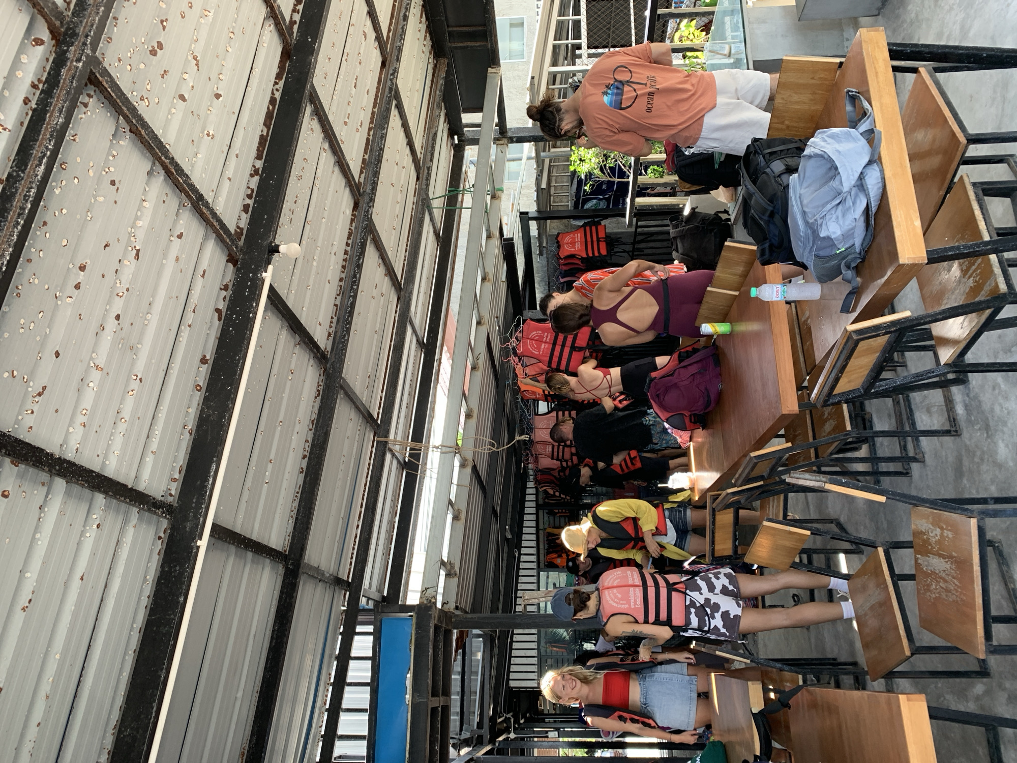 A group of tourists, dressed in casual wear and some wearing life vests, are gathered under a covered outdoor area. The structure appears to be a waiting area or staging point for an activity, possibly boat tours given the abundance of life vests. Tables and chairs are arranged throughout the space. The ceiling is made of corrugated metal with visible rust and staining, and there are exposed beams and lighting fixtures. In the background, there are buildings and tropical foliage, suggesting a location in a warm climate. The people are interacting casually, some are talking, and others are looking at their phones or waiting. The scene has a relaxed, informal atmosphere typical of tourist destinations. The overall impression is that of a group preparing for an excursion in a tropical location like Ko Samui District, Thailand.