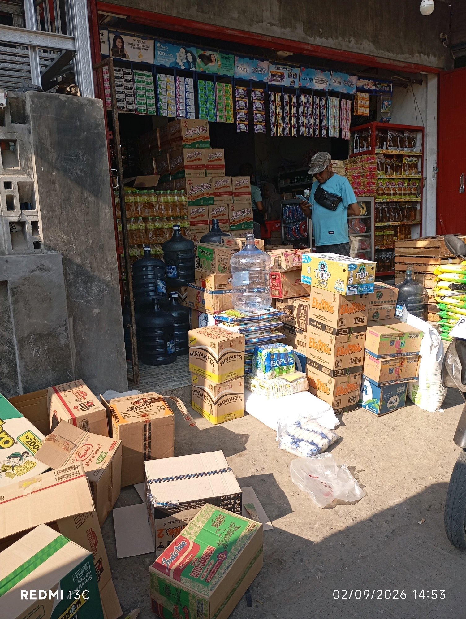 The image depicts a street-level view of a small shop or warung in Rambipuji, Indonesia. In the foreground, numerous cardboard boxes of various sizes are stacked and scattered on the concrete ground. Several boxes are identifiable by their labels, including "Indomie," "Sasa," "Top Coffee," and "Minyakita." Large blue plastic water jugs, likely for sale, are also present. A man wearing a camouflage cap, a light blue t-shirt, and a dark fanny pack is standing in front of the shop, looking at his phone. Behind him, shelves are stocked with packaged goods, including bottles of cooking oil and various other products. A partial view of the shop's interior reveals more shelves filled with merchandise. The overall scene suggests a commercial environment typical of a local Indonesian retail establishment. The lighting indicates it is daytime, with shadows cast by the objects and structures. A portion of a motorcycle is visible on the right side of the image. The timestamp "02/09/2026 14:53" is overlaid on the bottom right, and "REDMI 13C" is on the bottom left.