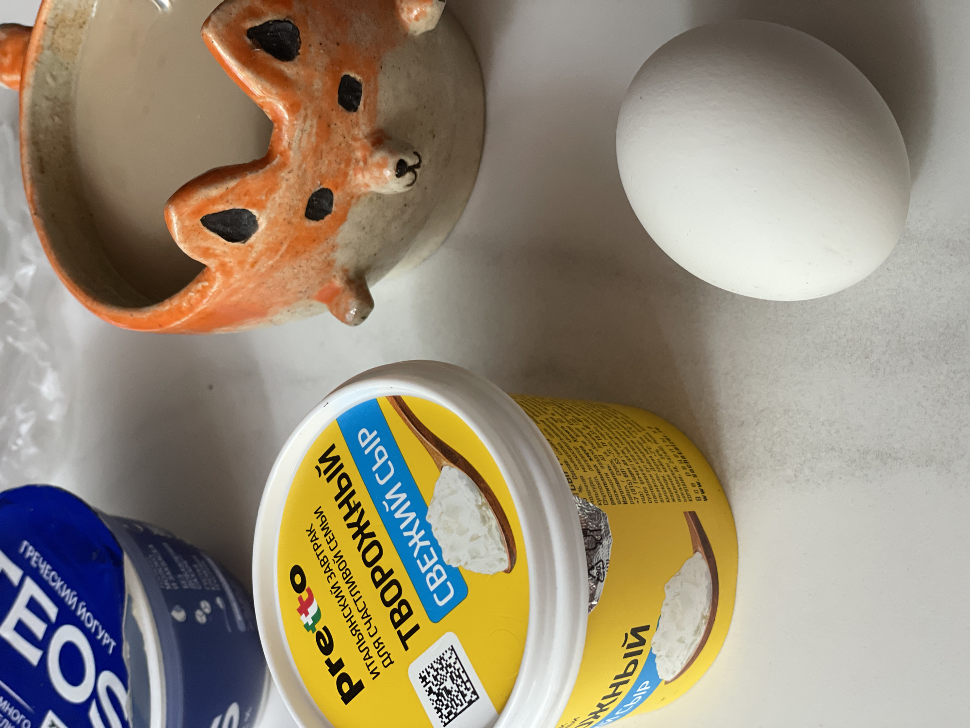 The image presents a flat lay arrangement of food items on a white surface. At the top left is a ceramic dish, possibly a planter, shaped like an animal, with orange and brown hues. To the right is a single white egg. Below these items, two tubs of dairy products are positioned. The tub to the left is blue with the brand name "TEOS". The second tub has a yellow label, with the brand name "preto" and text in Russian that translates to "cottage cheese". A QR code is also printed on the label. The label also states "ITALIAN BREAKFAST FOR HAPPY FAMILY". The photograph seems to be taken indoors in a brightly lit environment. The image has no visible people, actions, or interactions. The scene appears to be set on a table or countertop. Based on the OCR text that is visible on the yellow tub, a company located in Zheleznogorsk is mentioned ("Компани АК. 242440 Рес."). However, without more information, a definitive identification of the exact location within the city isn't possible.