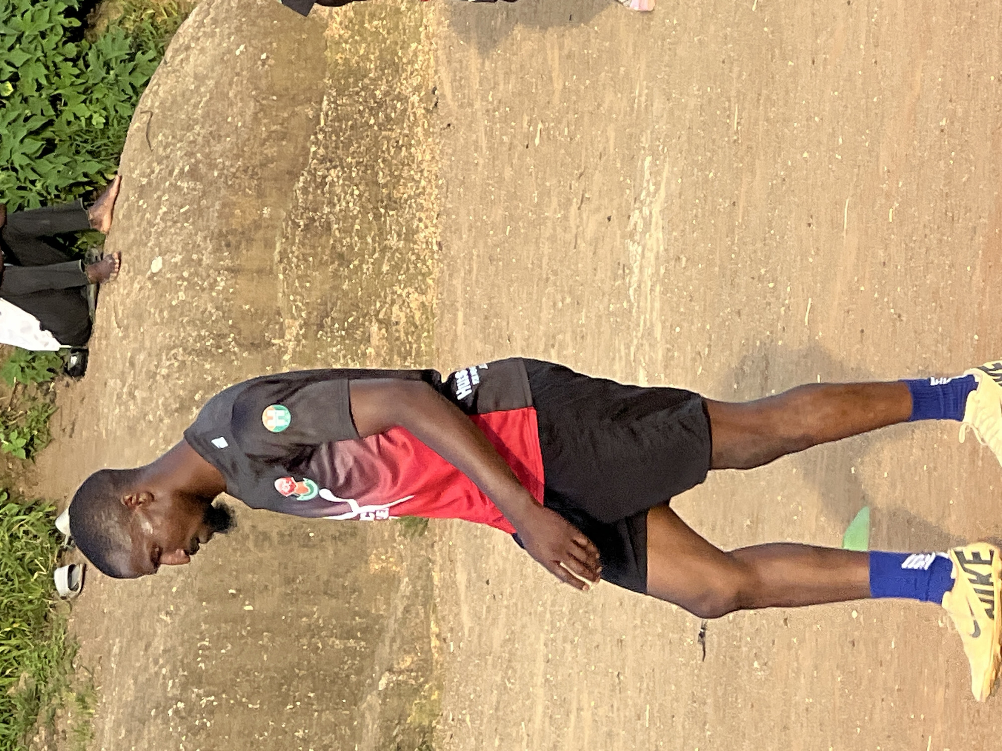 In the city of Jos, Nigeria, a Black man, appearing to be in his late 20s or early 30s, walks across a dusty, outdoor field. He is wearing a red and black athletic jersey with a logo on the left chest, black athletic shorts, blue socks, and yellow Nike cleats. His head is turned to his left, and he has a short beard. To the left of the man, a bright green cone is visible on the ground. In the background, a large, weathered rock formation is present, and on top of it, two people are seated, one wearing black pants and the other obscured. Green foliage is visible behind the rock.