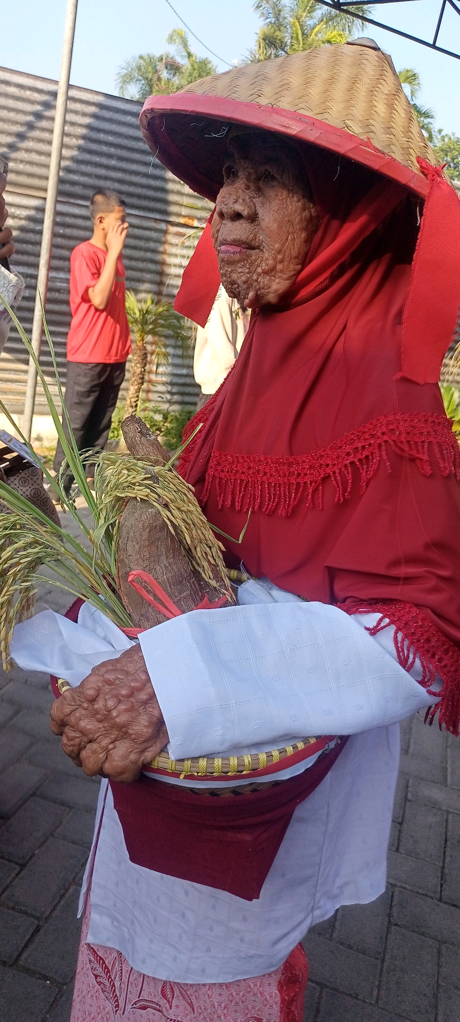 The image shows an elderly woman wearing a conical woven hat with a red brim and a red headscarf. She is also wearing a red garment with red fringe trim over a white garment and a patterned skirt. The woman is holding a basket containing a large piece of root or wood and stalks of rice. Her hands appear wrinkled and textured. In the background, a young boy in a red t-shirt and black pants is visible, as well as a palm tree and a corrugated metal fence. The scene appears to be outdoors, with a paved ground surface. The image was taken in Gempolklutuk, Indonesia.