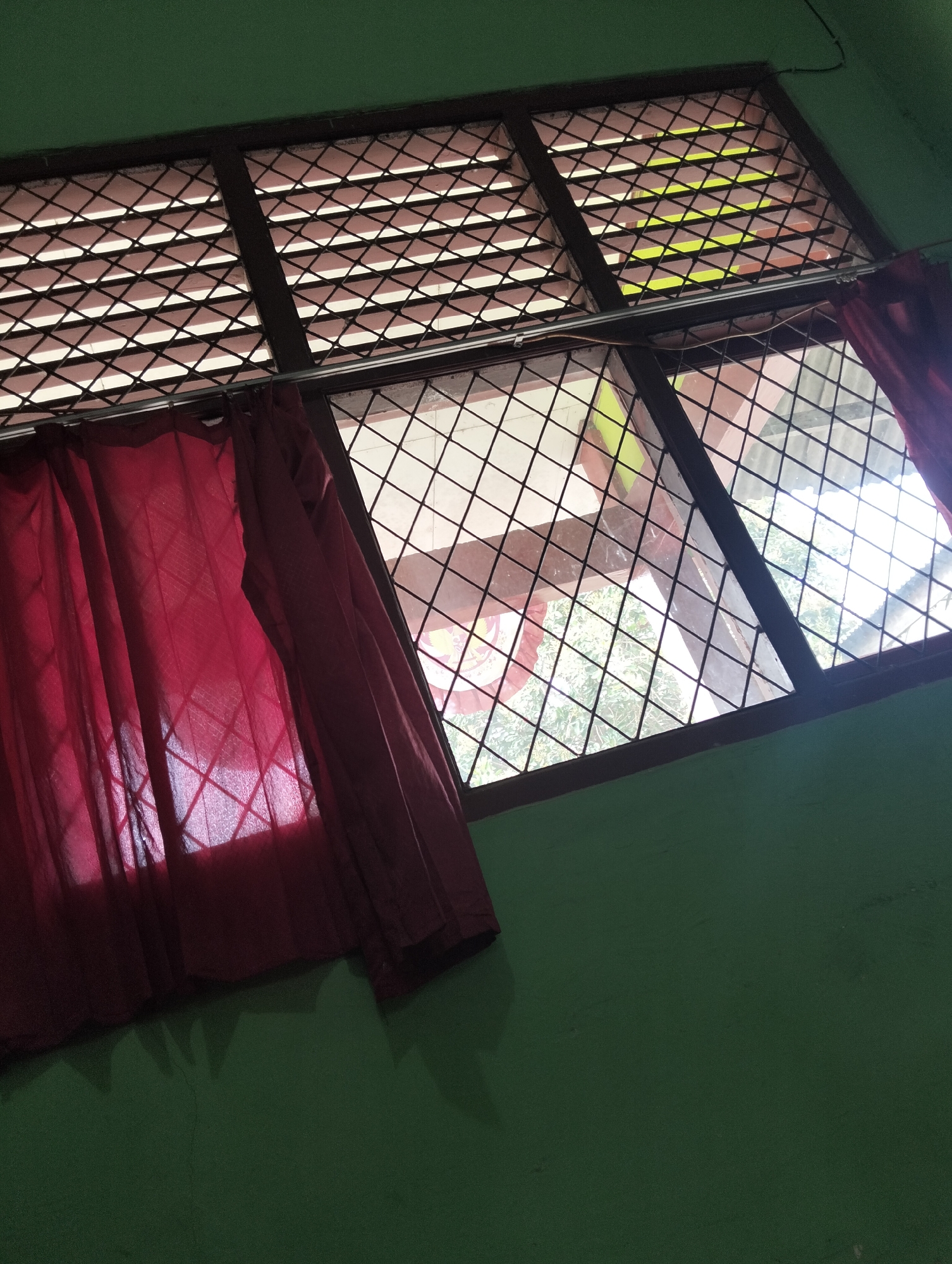 The image shows a window with a metal grate, covered partially by a red curtain. Outside the window, there are trees and sunlight visible. The wall next to the window is painted green. The scene appears to be indoors, looking out. There are no people or actions depicted in the image. The environment is domestic. The location is specified as Banjar Agung, Indonesia.
