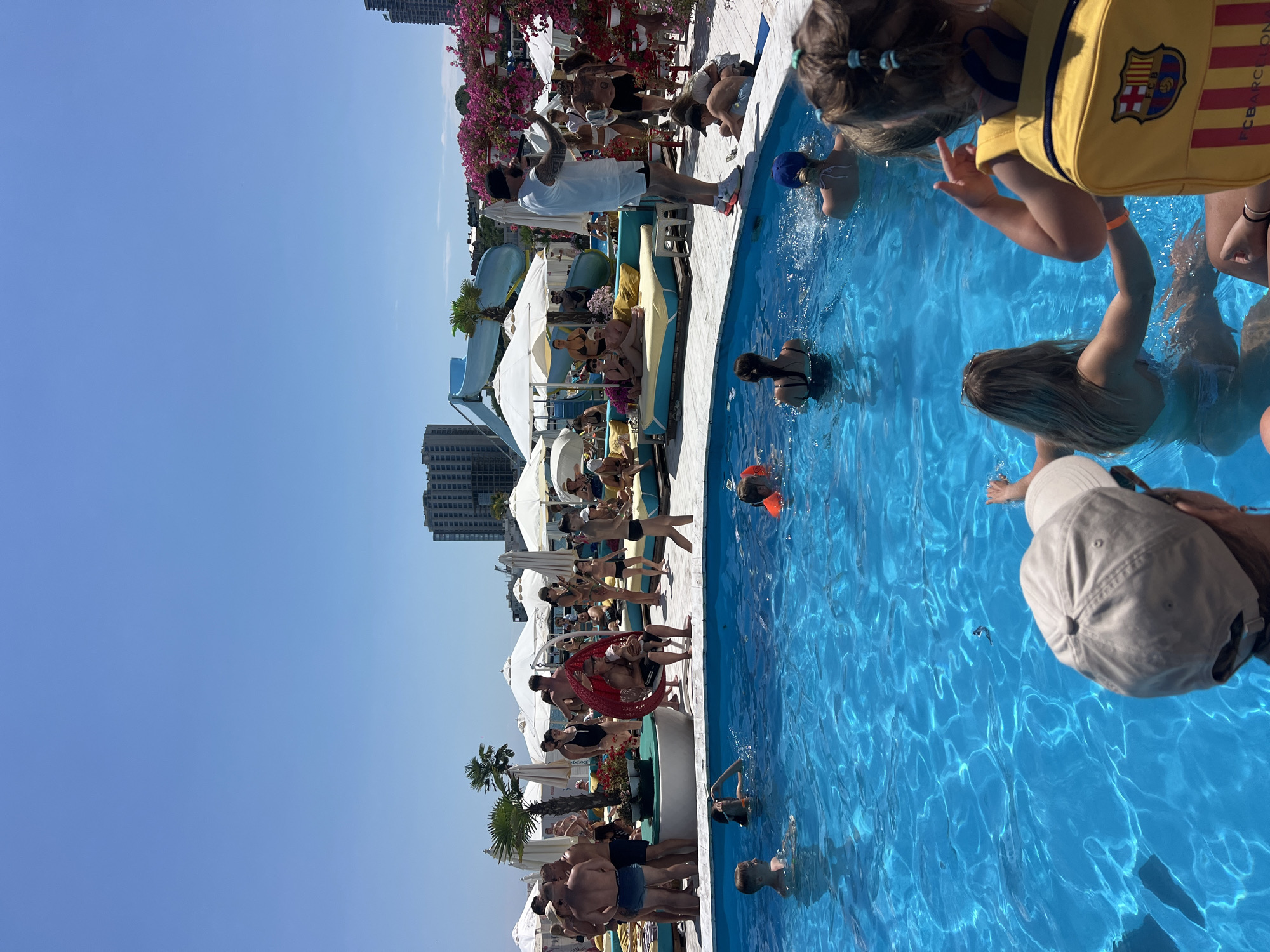 This is a scene at a pool party in Odesa, Ukraine, on a sunny day. The pool is a vibrant blue, with people of various ages swimming and relaxing. Some are in the water, while others are lounging on chairs and daybeds around the pool. There are palm trees and umbrellas providing shade. In the background, there are buildings and a water slide. A man is standing near the edge of the pool, holding a microphone, possibly entertaining the crowd. The atmosphere appears lively and summery, with many people enjoying the recreational facilities.