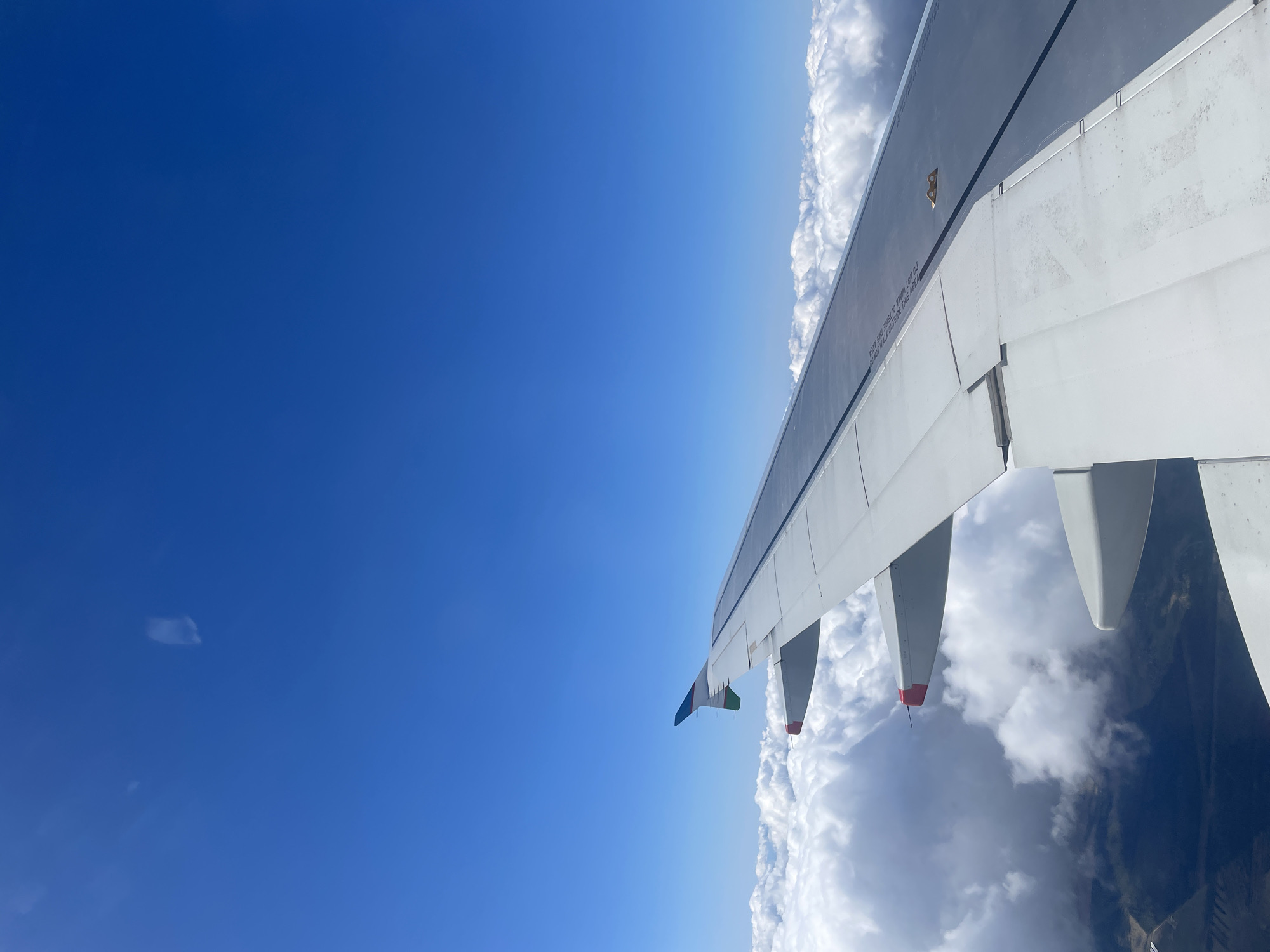 An aerial photograph, taken from an airplane window, shows the plane's wing against a backdrop of blue sky and clouds. The wing, primarily white with some gray and black components, extends diagonally across the frame from the upper right to the lower left. The leading edge of the wing is visible, along with several flaps in the extended position. The sky on the left of the frame is a clear, bright blue, contrasting with the fluffy white clouds visible on the right side, below the wing. Ground features are not visible in detail through the clouds. The image does not reveal any specific information that could place the scene in Saint Petersburg, Russia.