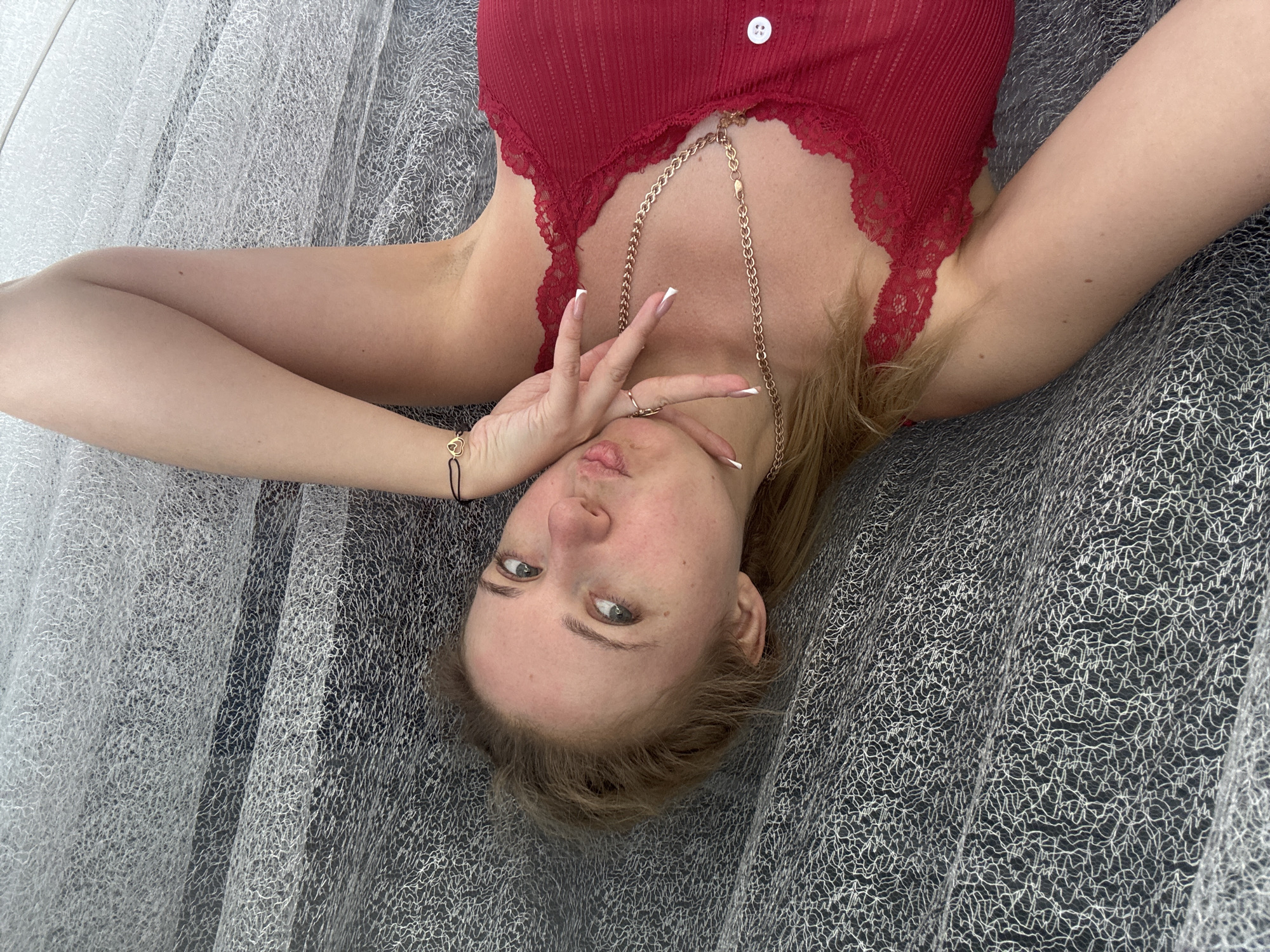 In Makiivka, Ukraine, a young woman lies down, propping her head with her left hand. Her right arm rests on her chest, displaying a gold chain necklace and a ring. She wears a red lace-trimmed tank top. The background is a textured grey surface. The scene appears to be indoors, possibly in a bedroom.