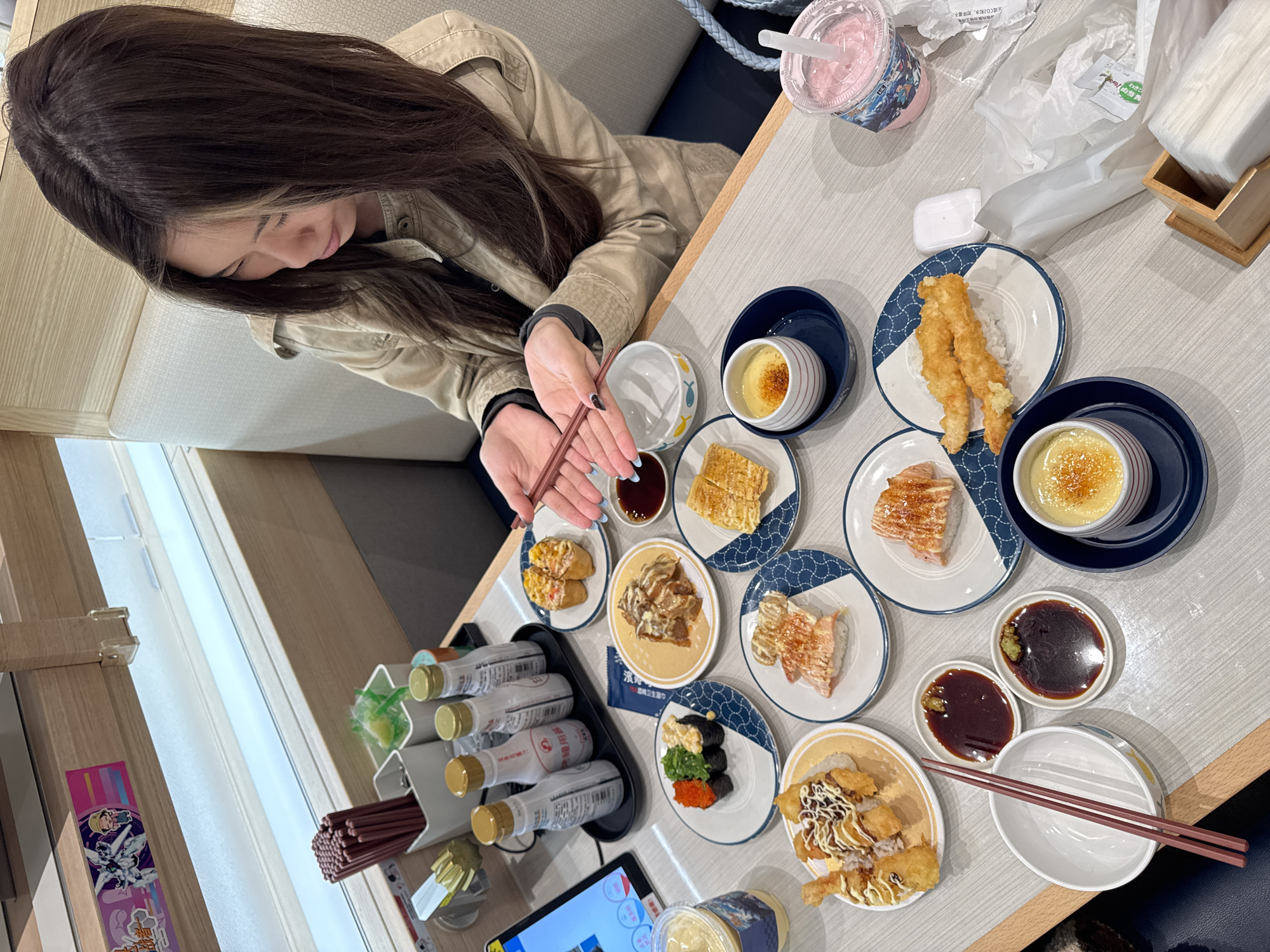 A woman with long brown hair sits at a table laden with food, likely in a restaurant in the Nan'an District of China. She is holding chopsticks, possibly about to eat. On the table are numerous small plates of sushi, tempura, and custard desserts, along with small bowls of soy sauce and condiments. There are also beverages in bottles and cups, along with napkins and a tablet-like device. The setting appears to be a brightly lit, modern restaurant.