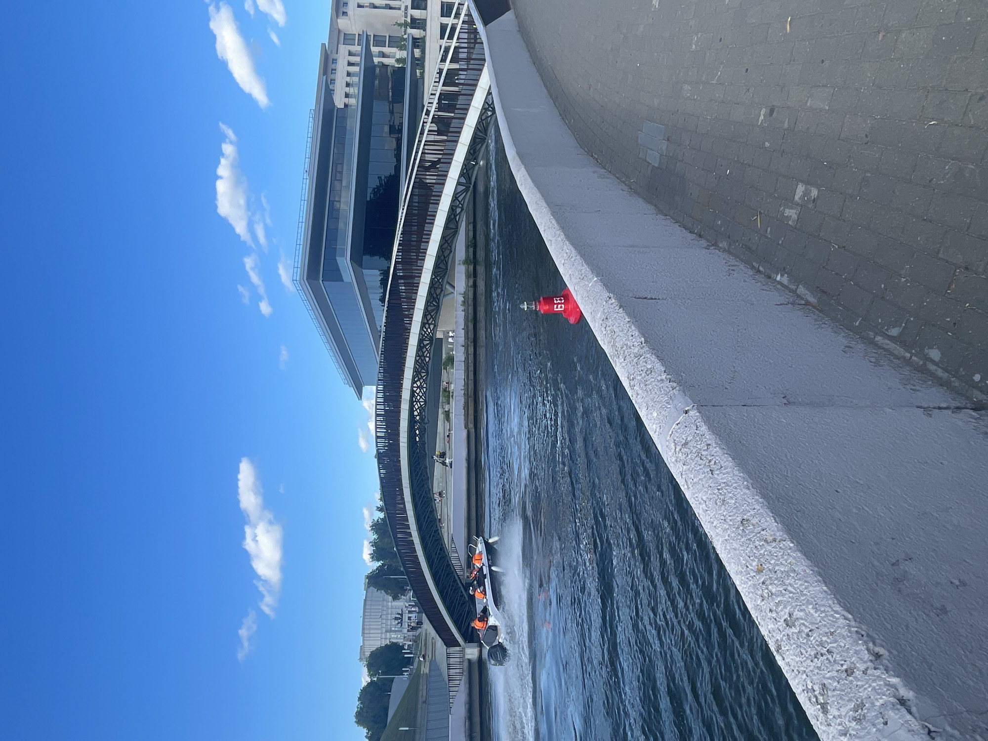 A white boat with several people wearing orange life vests is moving through the water, creating a spray behind it. The boat is seen from an elevated perspective, with the water appearing dark and rippling. In the background, a modern bridge with a metallic structure curves over the water. Beyond the bridge, there is a large building with glass windows and some greenery. The sky is a vibrant blue with a few scattered white clouds. A red buoy with white markings, including what appears to be the number "69", is visible in the water near the edge of the scene. The overall scene suggests an outdoor activity, possibly a boat tour or a watersport, taking place on a sunny day. The architecture of the buildings and the design of the bridge hint at an urban setting, perhaps a city with a waterfront.