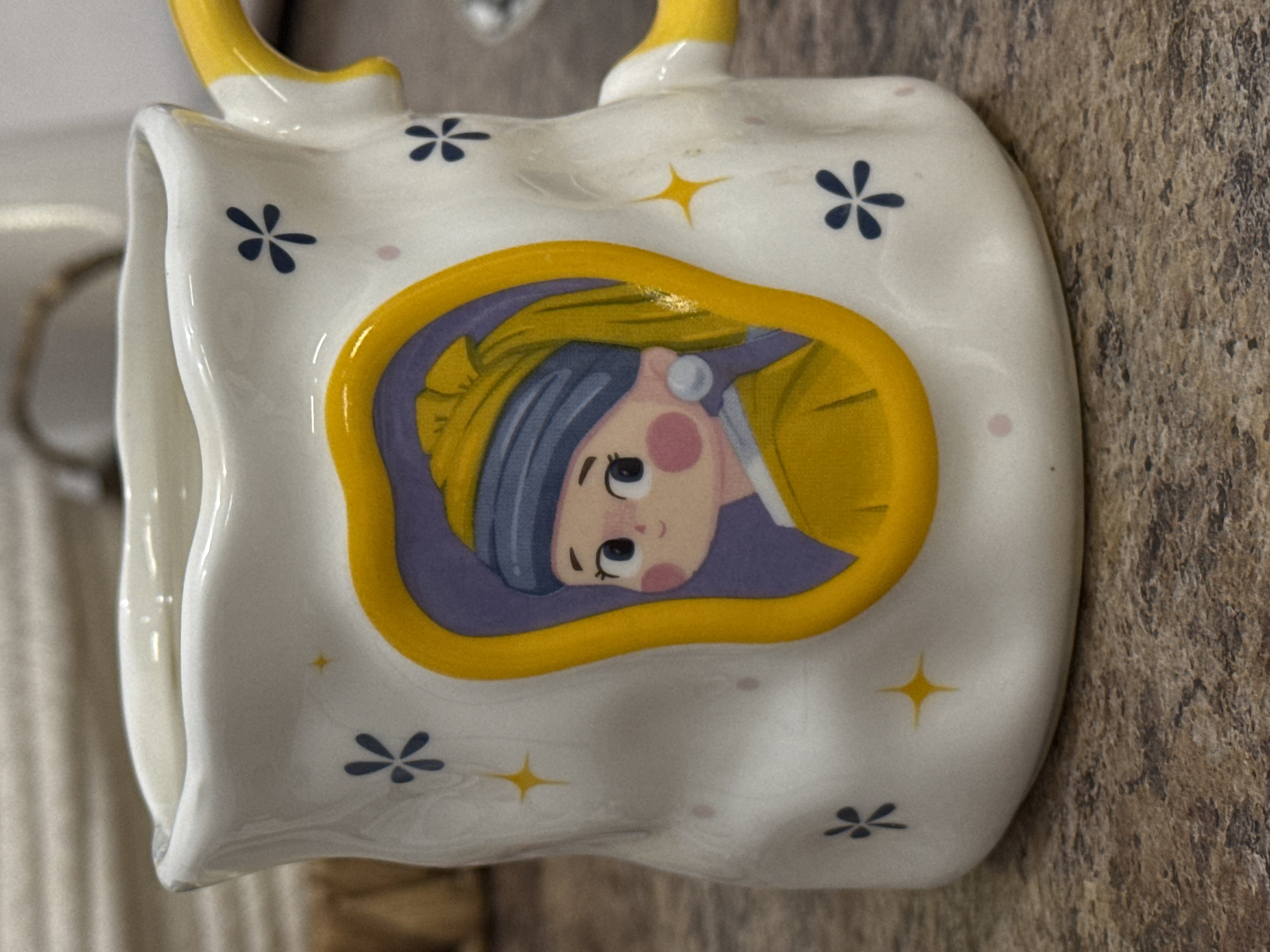 A close-up, eye-level shot shows a white mug with a yellow handle decorated with a cartoon image of a girl with a pearl earring. The girl is wearing a blue headscarf and a yellow outfit, and her portrait is framed in a yellow, irregular shape. The mug itself is adorned with small blue floral designs and yellow starbursts. The mug sits on a brown, speckled countertop. The background is blurred, showing vertical blinds or a similar textured surface. The overall scene is indoors.