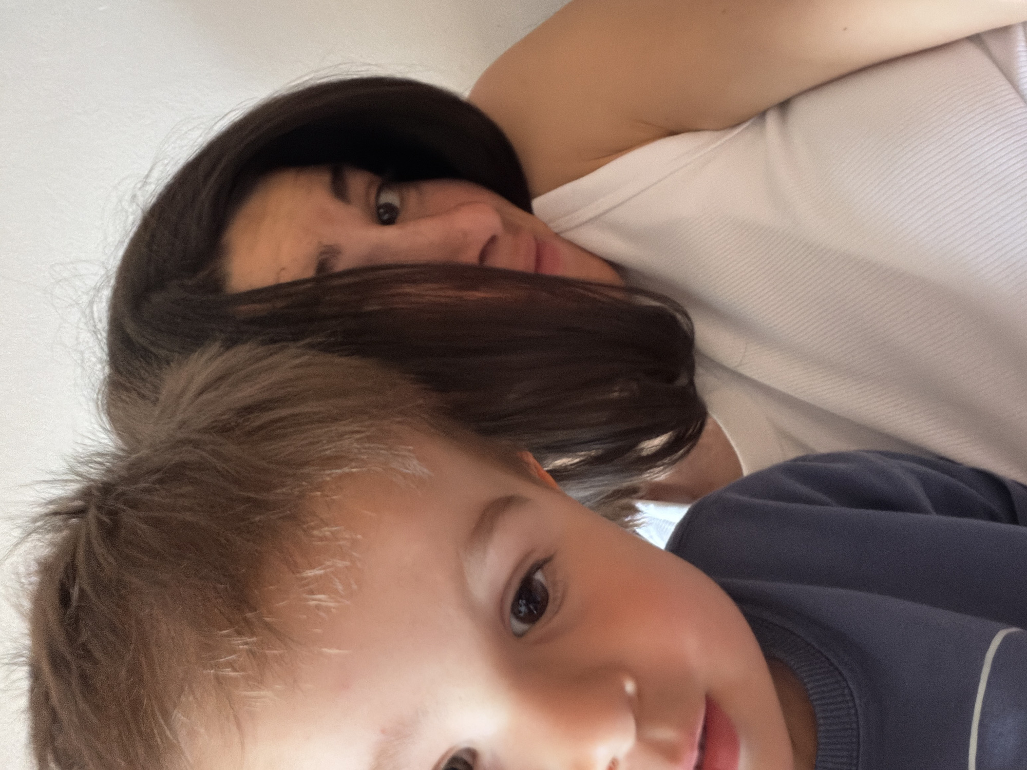 A selfie-style close-up shows a woman and a young child. The woman has dark hair partially obscuring her face, and she wears a white ribbed tank top. The child has light brown hair and wears a blue long-sleeved shirt with a white stripe on the sleeve. The background appears to be a plain white wall. The location in Budva, Montenegro is unknown.