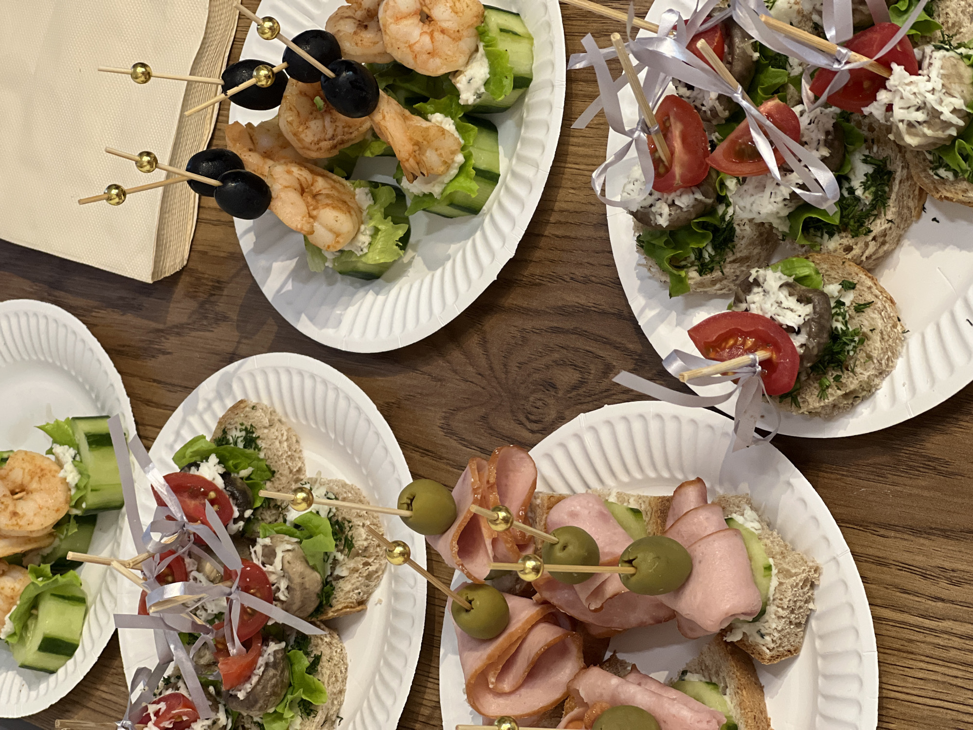 The image displays an overhead, close-up view of various appetizers arranged on white, corrugated paper plates, set against a wooden table. The scene suggests a catering event or a casual gathering.
In the upper left, a plate features skewers with cooked shrimp and black olives. These are presented on individual cucumber slices, each topped with a creamy white spread, likely feta or cream cheese, and a small piece of lettuce. Nearby, a stack of folded beige paper napkins is visible.
Another plate, located in the upper right, holds several open-faced sandwiches or canapés. These are made with slices of brown bread, topped with lettuce, a shredded white ingredient (possibly cheese), small dark pieces (appearing to be mushrooms or a similar ingredient), and fresh dill. Each canapé is garnished with a half cherry tomato and secured with a wooden skewer tied with silver ribbons.
In the lower right, a third plate showcases more open-faced sandwiches. These feature brown bread layered with thin cucumber slices, a white spread, and folded slices of light pink cold cuts, resembling ham. Each is topped with a green olive, held in place by a decorative wooden skewer with a gold spherical head.
The appetizers are neatly arranged and appear ready for consumption. No people or visible text are present in the image. The lighting is bright and even, typical of an indoor setting.