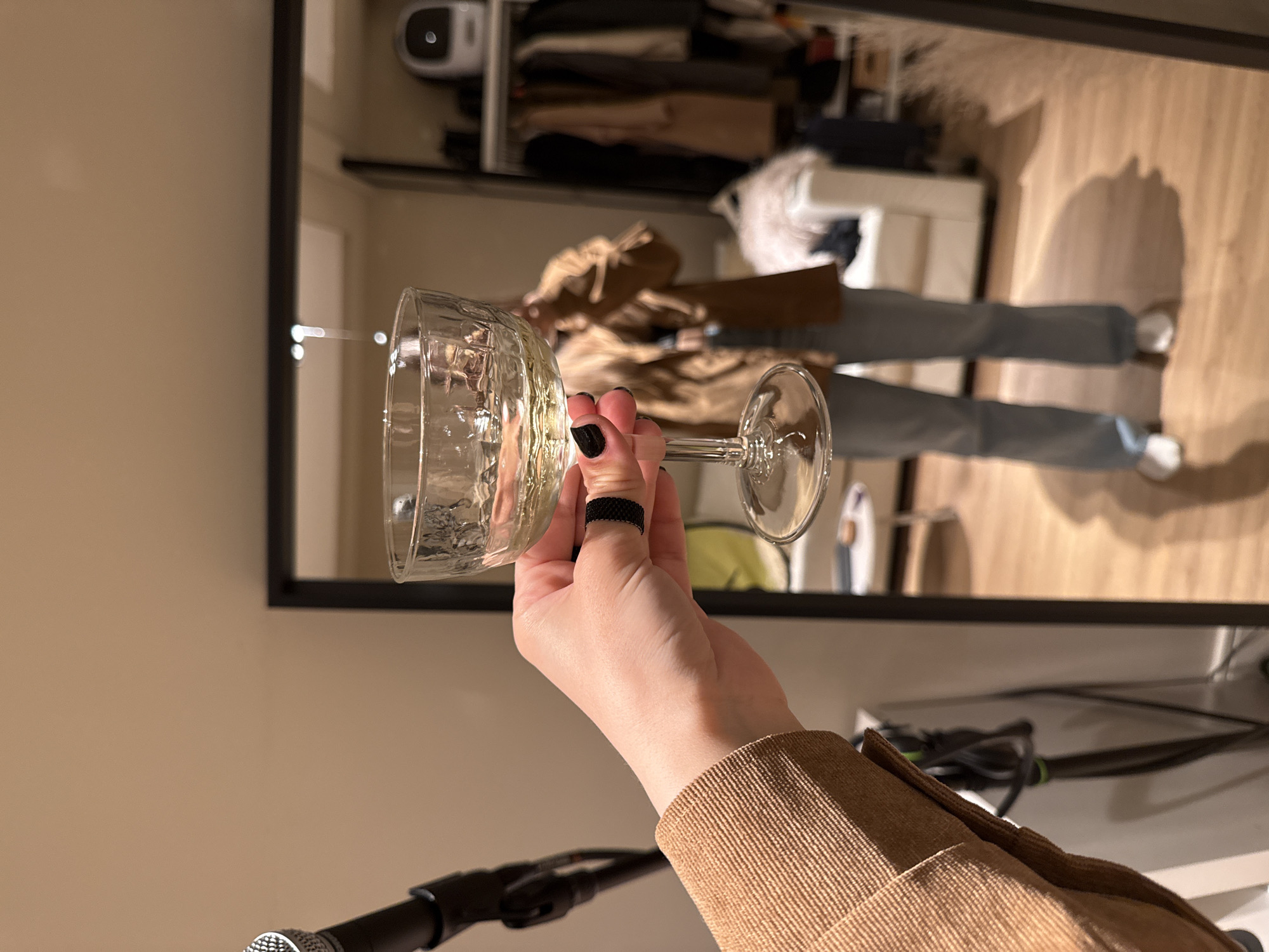 A hand holding a clear glass is positioned in front of a framed mirror, with a black microphone stand partially visible at the bottom. The hand has a black ring on one finger and black nail polish. The reflection in the mirror reveals a person wearing jeans and a tan jacket or shirt, standing in what appears to be a closet or dressing area. The background contains clothing and other personal items. The environment is indoor, with a warm lighting. There are no indications that the location is in Barcelona, Spain.