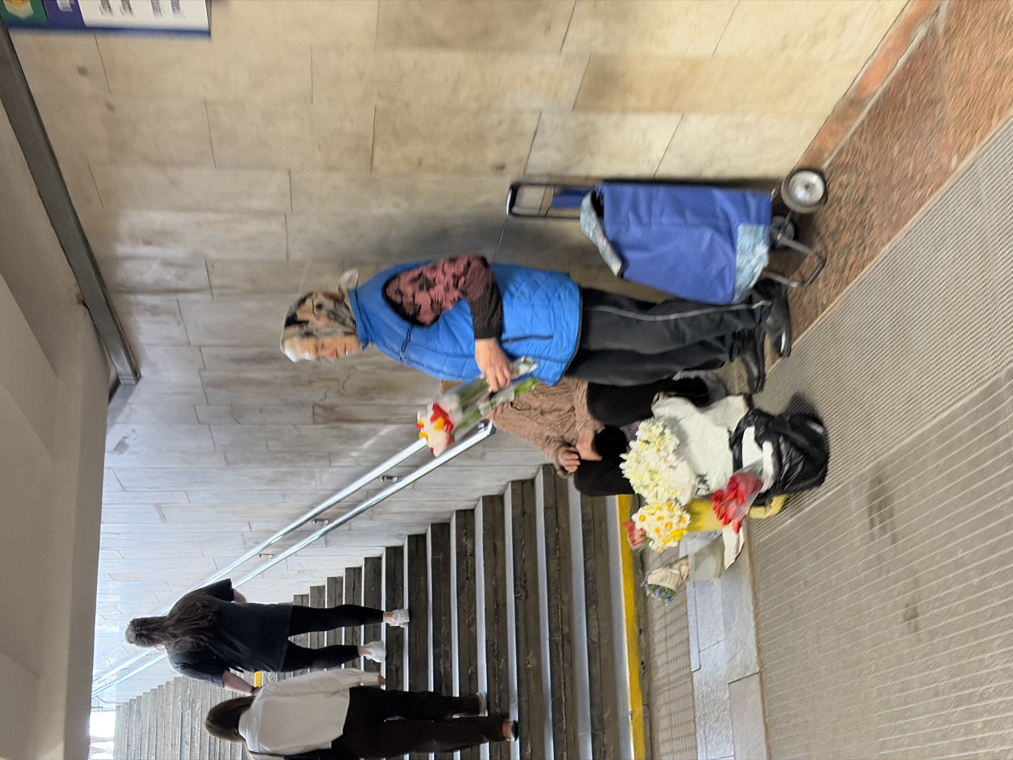 A woman in a blue vest and patterned headscarf stands on the sidewalk next to a set of stairs, holding a bouquet of flowers and with more flowers in a shopping cart and bag beside her. Two other individuals are walking down the stairs. The location appears to be an urban underpass in the Бараўлянскі сельскі Савет, Belarus. The environment consists of tiled walls, stairs with metal railings, and a concrete sidewalk with a textured finish.