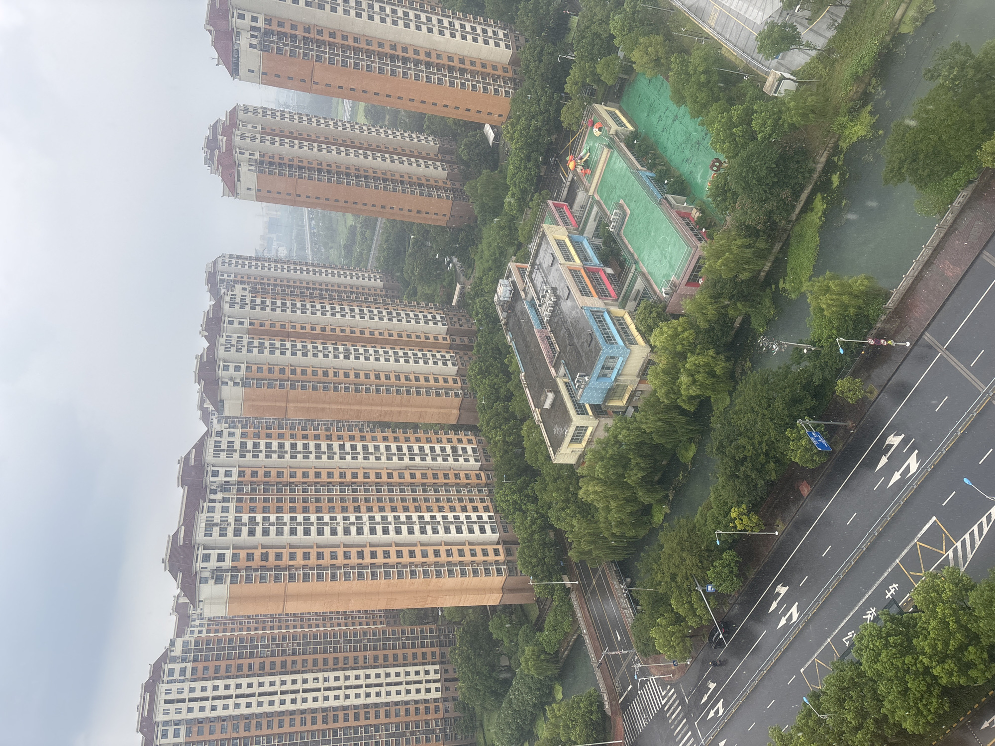 This is an aerial, eye-level, wide shot of a rainy day in the Suzhou Industrial Park, China. A row of tall, multi-story apartment buildings with orange and white facades and reddish-brown roofs are the dominant structures. In the foreground, a multi-lane road runs diagonally across the frame, wet from the rain, with white lane markings and arrows. Pedestrians are absent. A single black car is visible on the road. Adjacent to the road is a canal or river, flanked by lush green trees, some of which appear to be willow trees. Across the canal from the apartment buildings is a colorful, multi-story school building with a green playground area on its roof. The sky is overcast and a light rain is falling, visible as small streaks throughout the image.