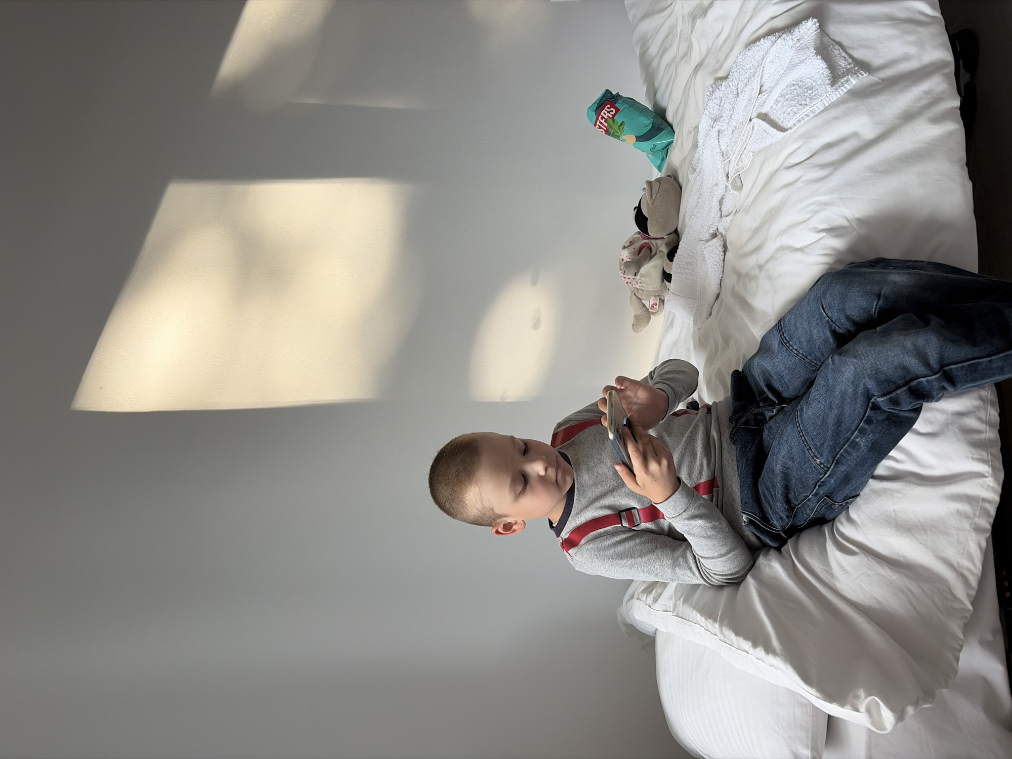 A young boy sits on a bed, engrossed in his smartphone. He is wearing a gray long-sleeved shirt with red suspenders and blue jeans. The bed is covered in white linens, and a white towel is neatly folded on the right side. Next to the towel are two stuffed animals and a bag of chips. Sunlight streams in from the right, casting a warm glow on the wall and creating shadows. The overall scene is peaceful and intimate, suggesting a moment of quiet leisure. The location is unknown.