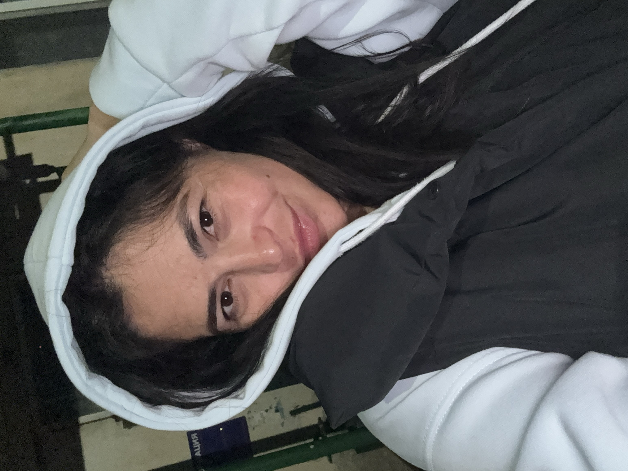 A woman with dark hair and brown eyes is wearing a white hoodie and a black jacket. She is looking directly at the camera and smiling slightly. The location appears to be indoors, possibly on bleachers, in the city of Кемуль, Russia.