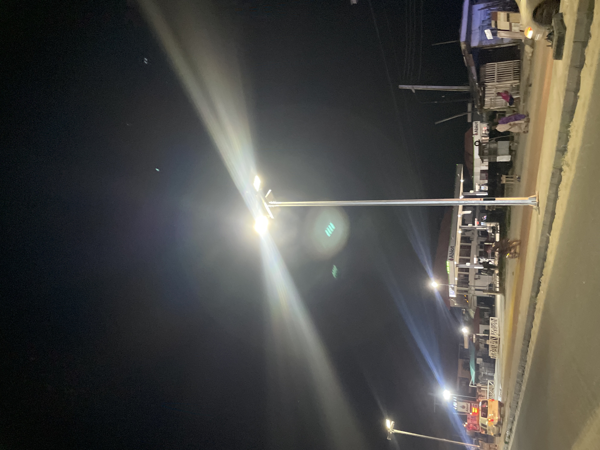 At night in Ado, Nigeria, a well-lit street is lined with businesses, including an "Ablot" branded building. People are visible on the street, and streetlights illuminate the scene.