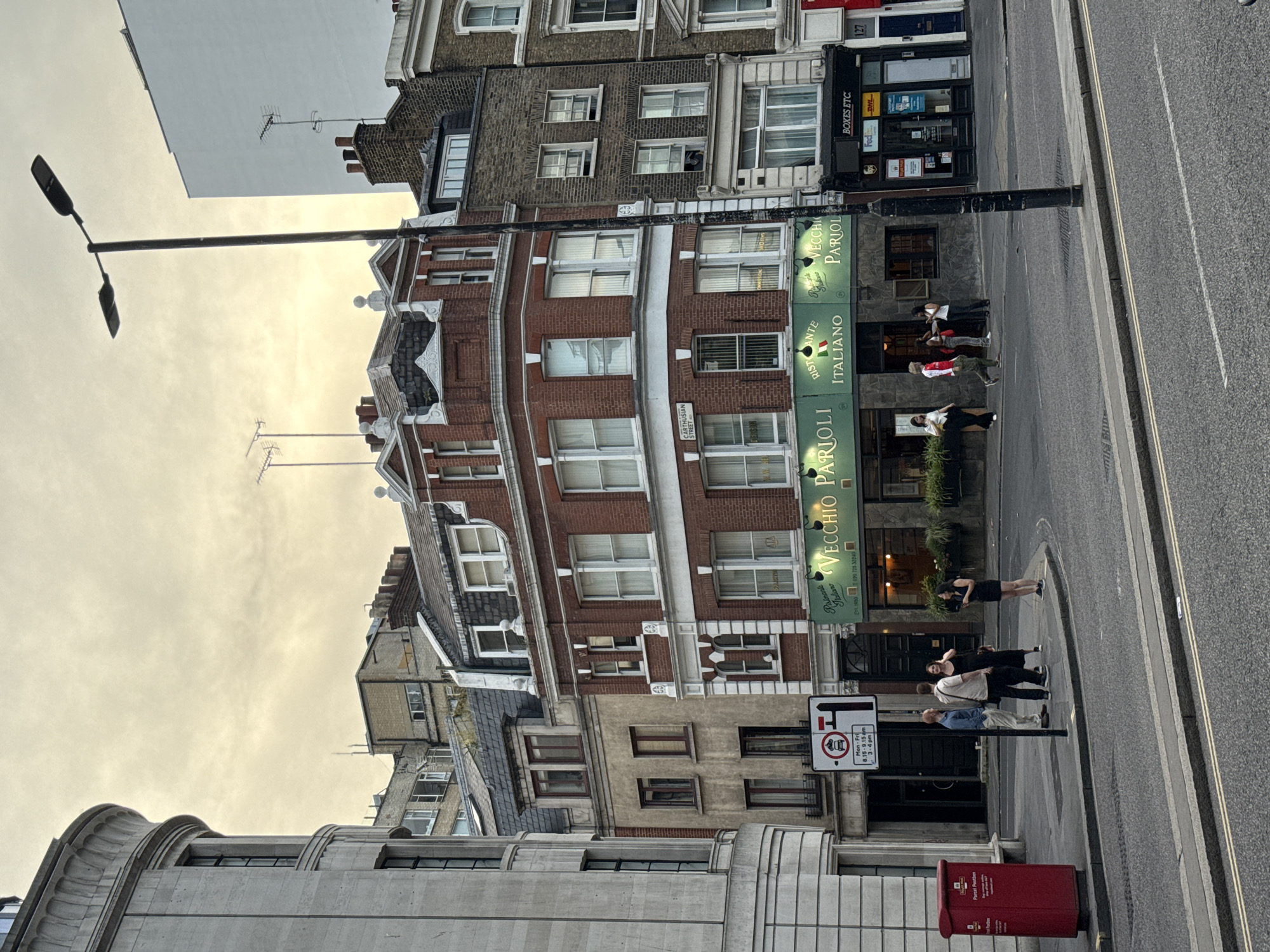 This is an outdoor daytime shot of a street in London, United Kingdom. The buildings are varied in architectural style. A sign reads "VECCHIO PARIOLI RISTORANTE ITALIANO." Another sign indicates "CARTHUSIAN STREET". People are on the pavement near the street. There is a red postal box and a street sign. The sky is overcast.