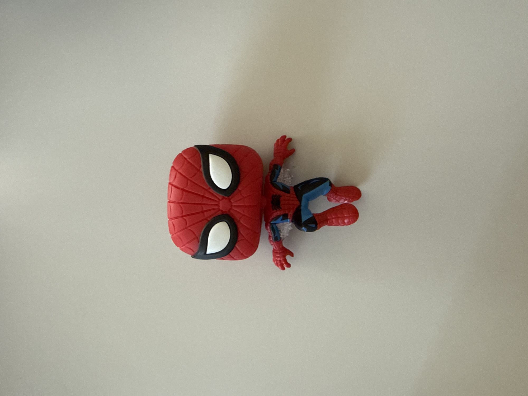 A Spiderman Funko Pop figurine is centered on a plain, off-white background. The figurine is predominantly red with black and blue accents, depicting Spiderman in a dynamic pose. Its head is large and round, characteristic of the Funko Pop style, with white eyes outlined in black. The body is smaller, with detailed representations of Spiderman's suit. The figurine appears to be standing or mid-action, with its arms outstretched. The lighting casts a subtle shadow to the right of the figurine. The environment does not provide any specific details that would allow for location identification within Bad Sachsa, Germany.