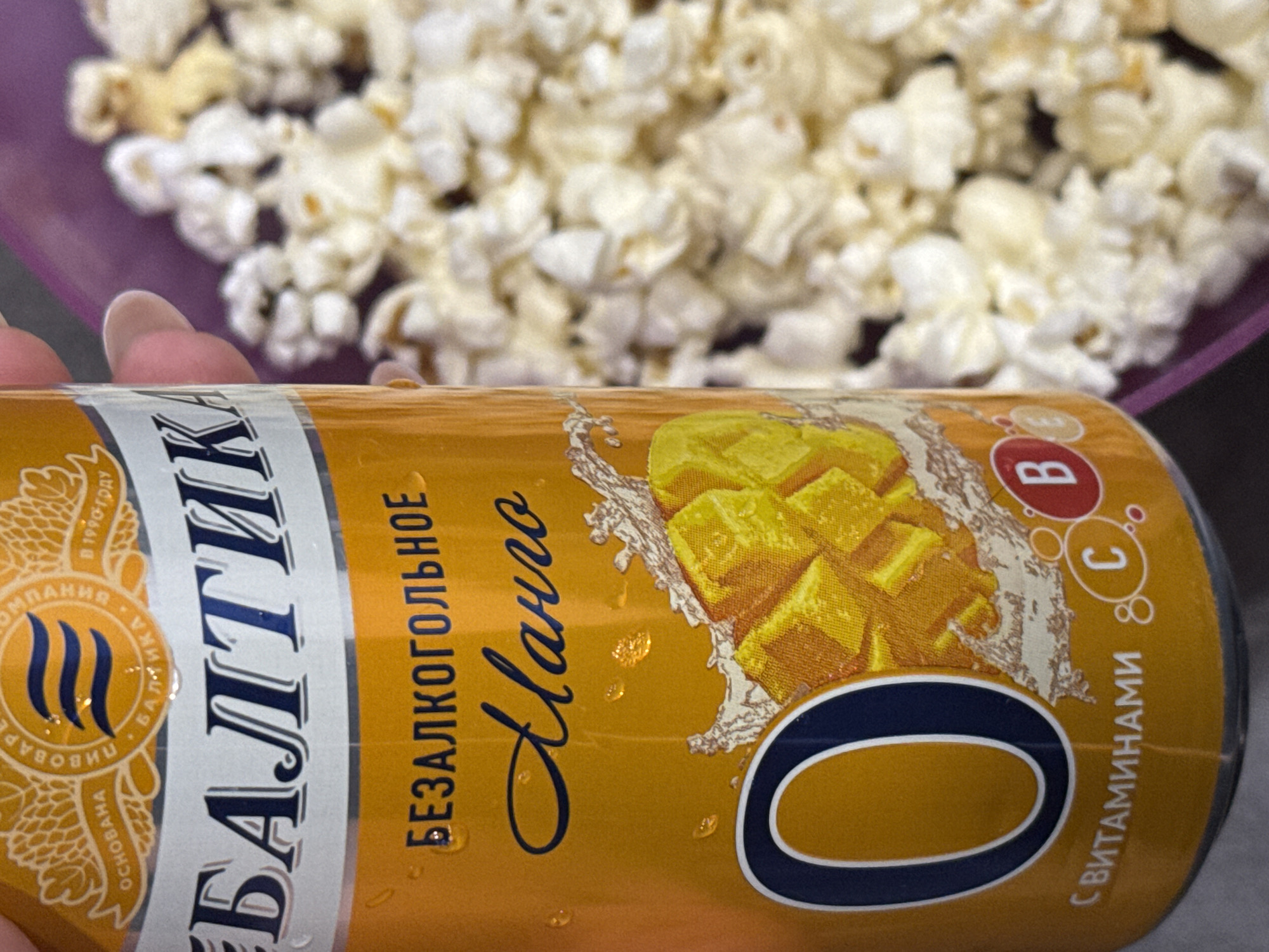 A can of Baltika 0 Mango, a non-alcoholic beer, is in the foreground. The can is predominantly gold with the brand name "Baltika" in large, dark blue Cyrillic letters. Below, the words "БЕЗАЛКОГОЛЬНОЕ" (non-alcoholic) and "Манго" (Mango) are also written in Cyrillic. An image of mango chunks with water splashes is on the can. A "0" logo is prominently displayed, and the words "С ВИТАМИНАМИ" (with vitamins) are present, along with the letters "B" and "C" circled.
In the background, there is a bowl of popcorn, out of focus. The scene is indoors, but the specific location is unknown. There are no people in the image except for a hint of fingers holding the can.