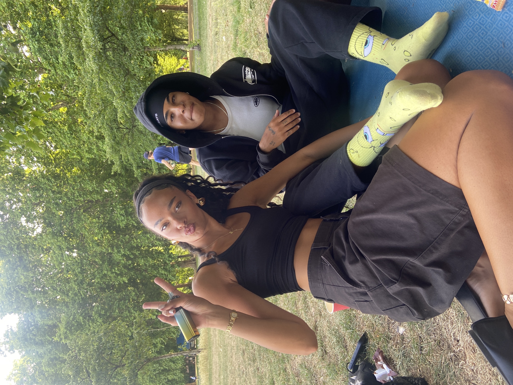 Two individuals are seated on a blue mat on a grassy area, with a backdrop of lush green trees. The person on the left, wearing a black tank top and dark shorts, is holding a metallic object in their raised hand, making a peace sign gesture. They have dark, curly hair and are looking towards the camera with pursed lips. A gold necklace and bracelet are visible. The person on the right is wearing a black hooded jacket over a white ribbed undershirt and black sweatpants. They have dark hair covered by a black headscarf and are also looking at the camera. Their feet are adorned with bright yellow socks featuring cartoon characters. The environment suggests a park setting.