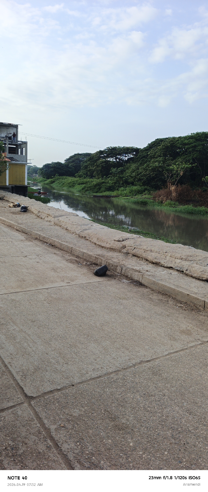 The image shows a river scene in Arismendi, Venezuela. A concrete walkway borders a muddy river lined with dense, green trees on the opposite bank. A partially constructed building stands to the left, with a person visible on the top floor. Debris lies on the walkway near the building. A black object, possibly a bag, sits on the walkway. The sky is overcast.