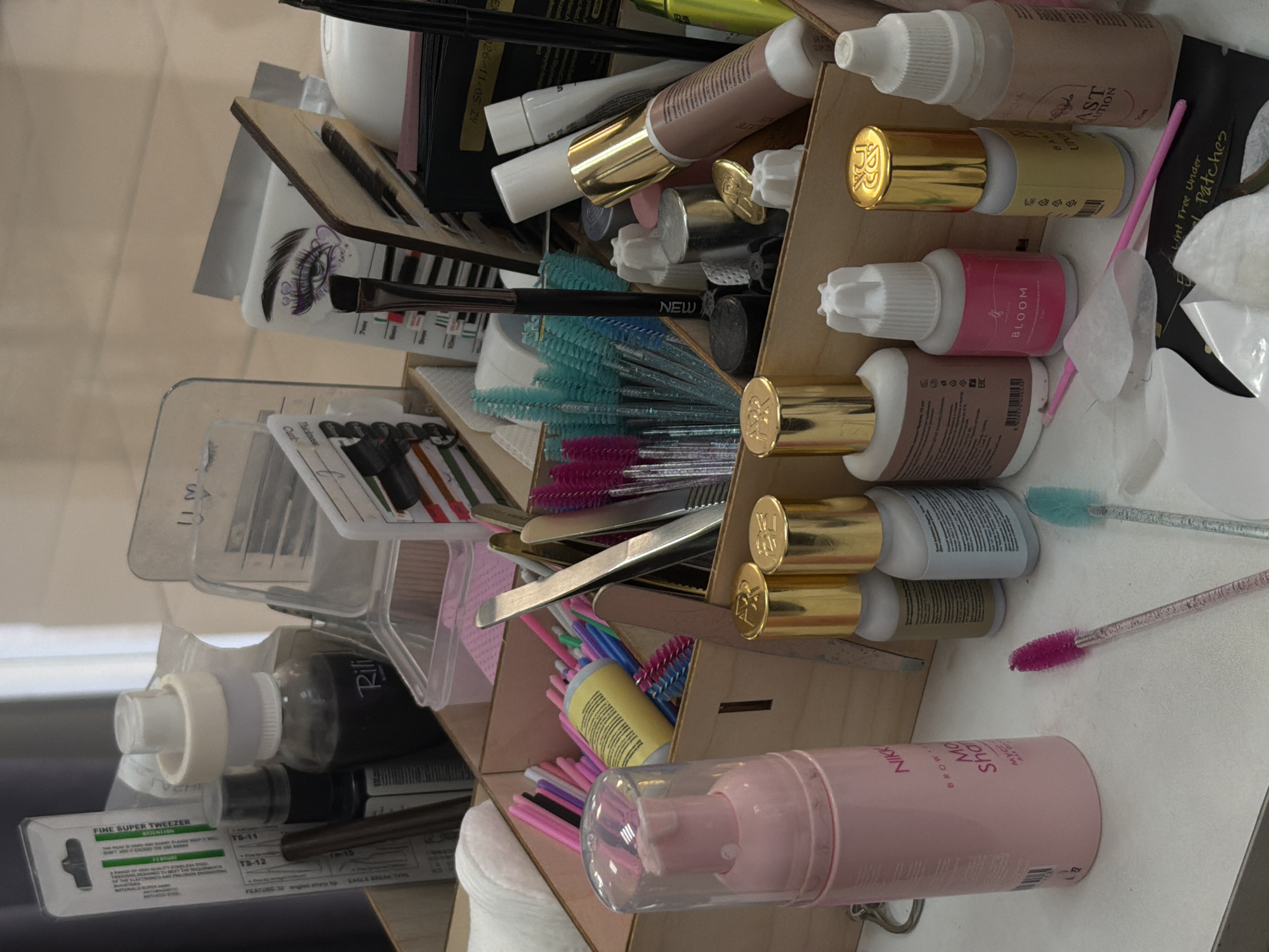 The image presents a close-up, overhead shot of a wooden organizer filled with various beauty and cosmetic supplies. Predominantly, there are multiple bottles with white caps and gold accents, some labeled with brand names such as "BLOOM," "NIKK," and "Rili." Also visible are eyelash extensions, tweezers, mascara wands in an array of colors, and lint-free under-eye patches. The environment appears to be a workspace, likely a beauty salon or personal vanity area. The specific location in Chaikovsky, Russia, cannot be determined from this image alone.