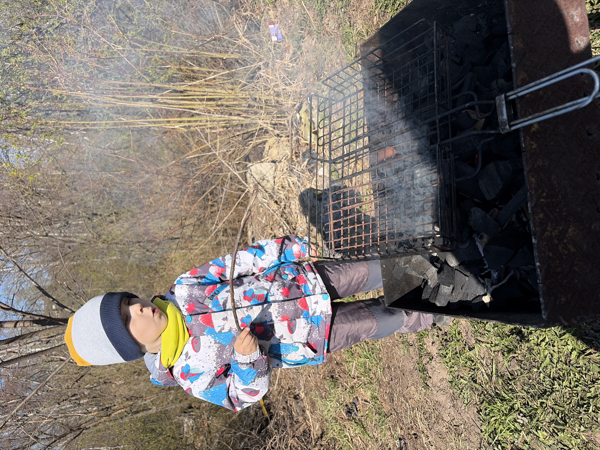 A child stands next to a charcoal grill in an outdoor area. The grill is filled with charcoal and topped with a metal grate. Smoke is visible around the grill. The child is wearing a colorful winter jacket, a yellow scarf, and a gray, white, and yellow beanie. They are holding a twig in their hand. The background consists of trees, bushes, and grass. Based on the prompt information, the location is Чумна, Russia.