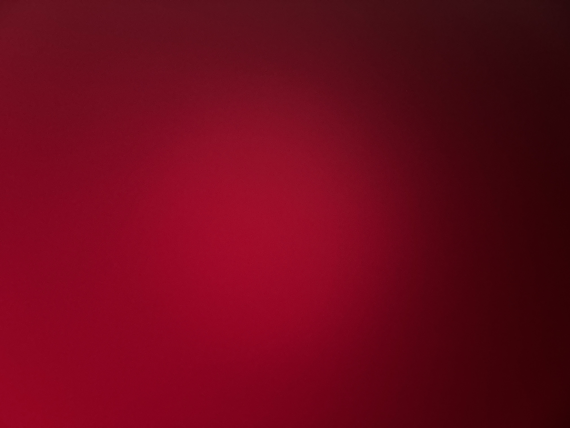 This is a horizontal, close-up, still photograph of a red surface that appears to be fabric. The lighting creates a gradient effect, with the color being a vibrant red on the left and fading to a deep maroon or almost black on the right. The texture of the surface is slightly grainy, suggesting a woven material. There are no people, objects, text, or discernible setting cues within the image. The overall impression is abstract, focusing solely on the color and texture of the red material. The abstract nature of the image, combined with its focus on a single color, makes it difficult to assign a specific time of day, weather, or location. The crops provided do not reveal any additional details about the subject matter.