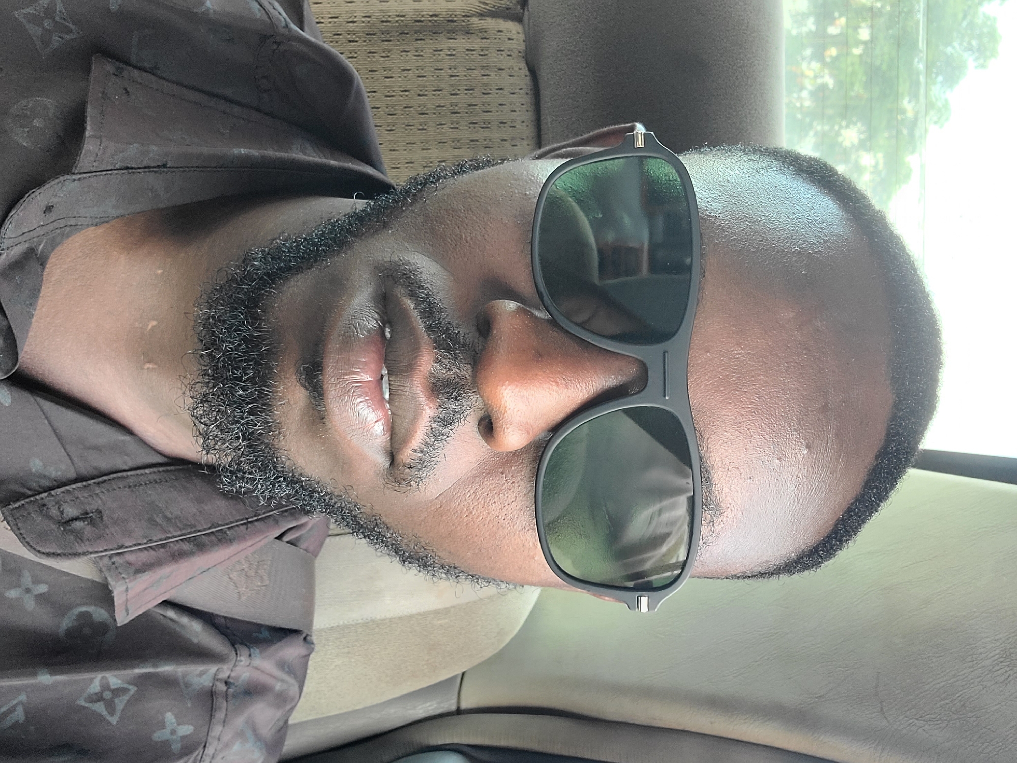A close-up selfie taken inside a vehicle shows a Black man wearing sunglasses and a dark brown shirt with a repeating pattern of stylized flowers. The man has short black hair and a full beard and mustache. He is seated in a vehicle with a light-colored headrest and a patterned seat visible behind him. The background through the window appears to be foliage.