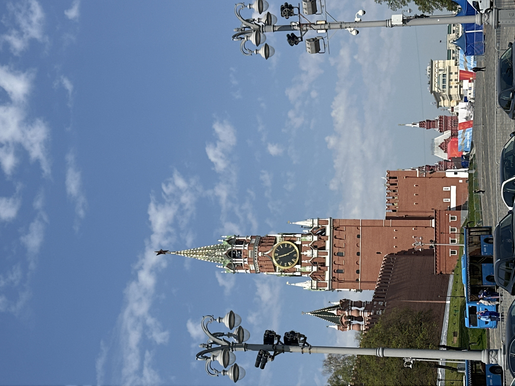 The image features a view of the Spasskaya Tower of the Moscow Kremlin in Moscow, Russia. The tower is a prominent red brick structure with a clock face and a red star on top. The sky is blue with scattered clouds. In the foreground, there are streetlights and what appear to be parade vehicles or maintenance carts lined up along a street. Some pedestrians are visible in the background, along with other buildings that are part of the Kremlin complex and other structures in the vicinity.
