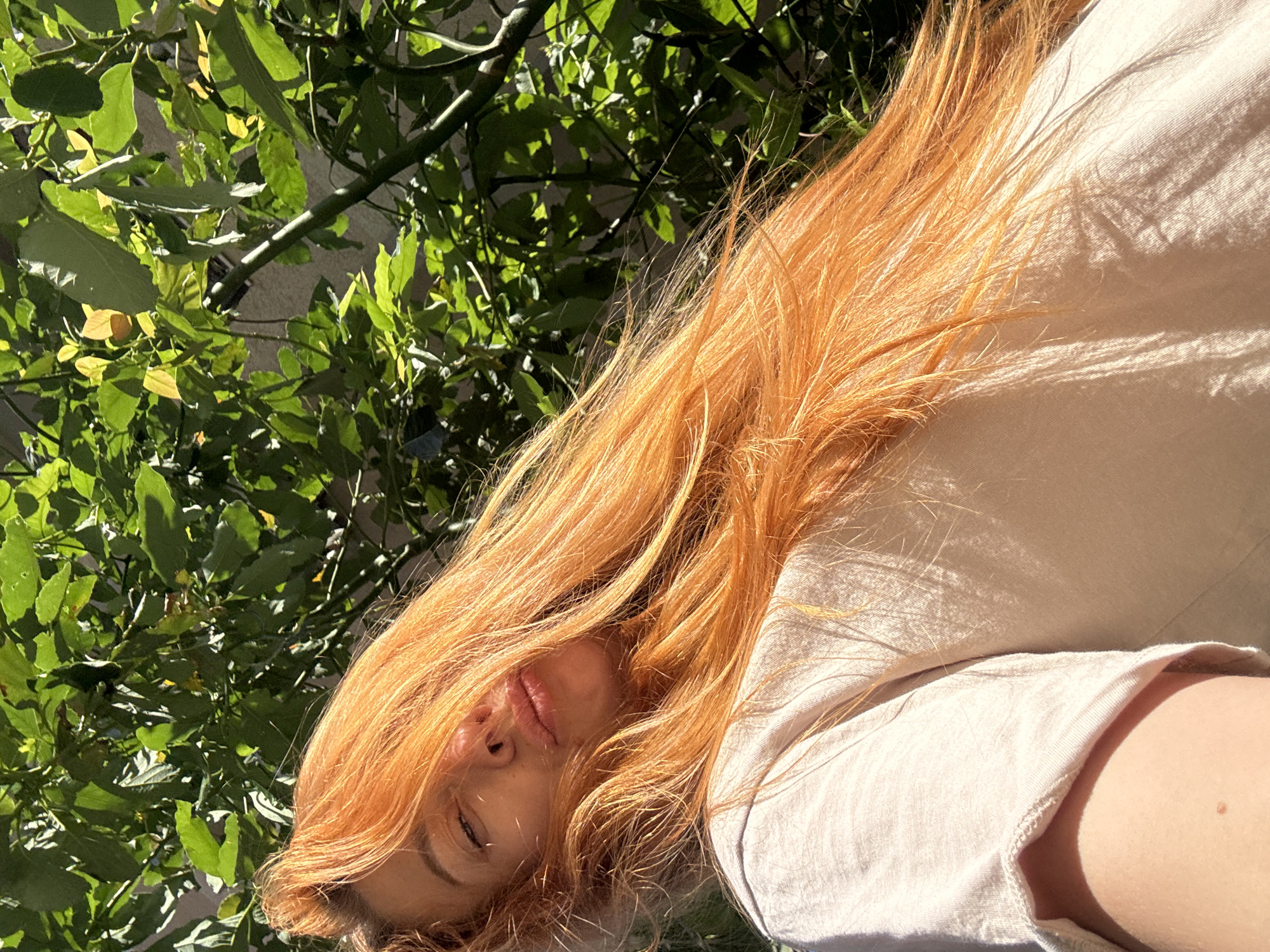 This is a close-up, low-angle selfie of a young woman with long, fiery orange hair. She is lying down outdoors, with lush green foliage partially obscuring the background. The sun is shining brightly, casting dappled shadows on her clothing and hair, indicating it's likely daytime. She is wearing a light-colored, possibly linen, t-shirt. Her eyes are closed, and her lips are slightly parted, suggesting a relaxed or peaceful state. The vibrant green leaves and the warm sunlight evoke a sense of nature and warmth. The location context provided is Budva, Montenegro, which is a coastal town known for its beaches and natural beauty, aligning with the outdoor scene. There is no visible text in the image.