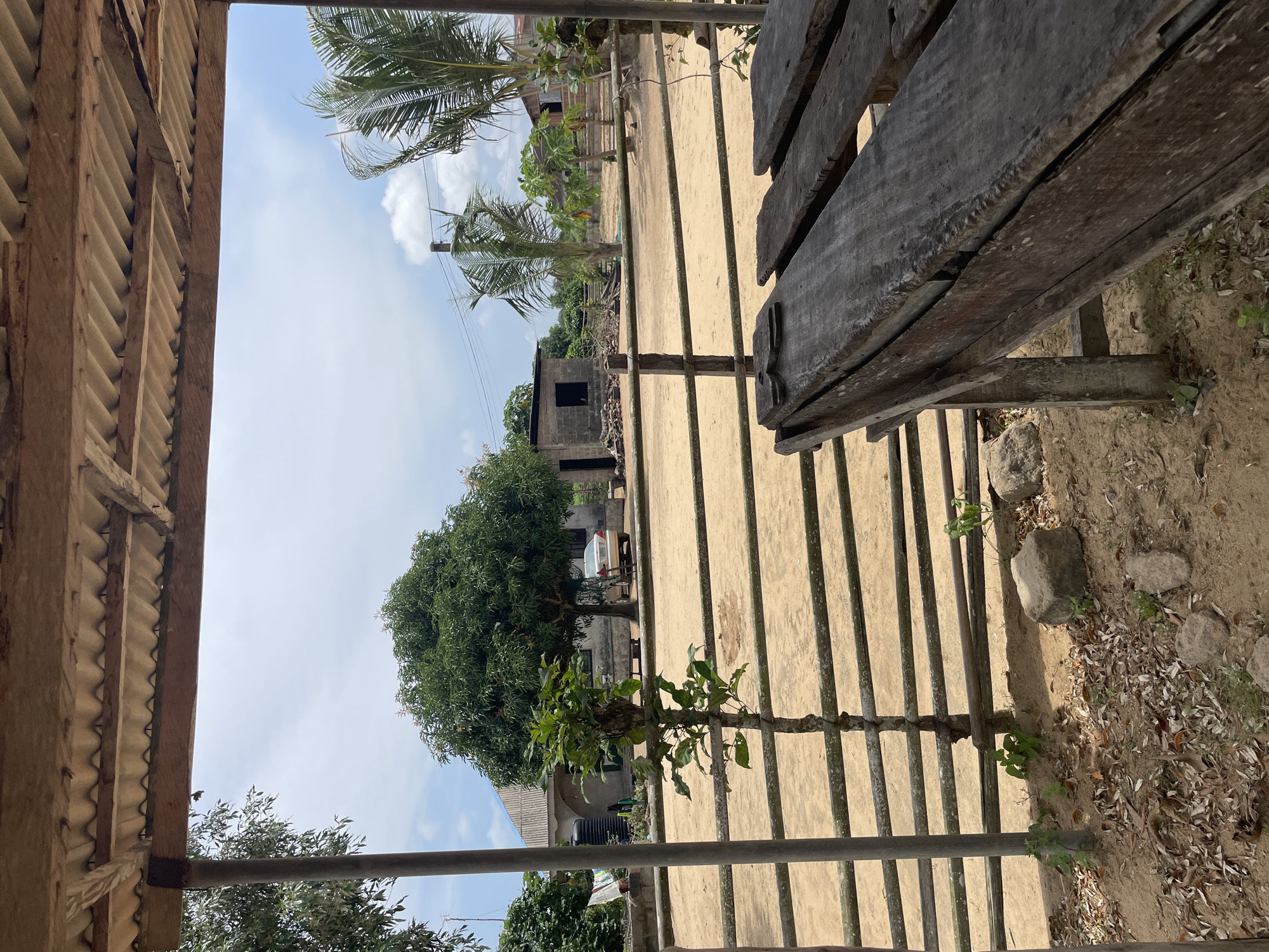 The image depicts an outdoor scene in Iyafin, Nigeria. In the foreground, a weathered wooden table and benches are partially visible, suggesting a rustic outdoor seating area. Beyond the table, a dirt yard is enclosed by a simple wooden fence. A small tree grows in front of the fence, and further back, a larger tree stands prominently. In the middle ground, a light-colored car is parked near a building with a corrugated roof. Another smaller, brick structure with a dark opening is also visible. Palm trees and other vegetation are scattered throughout the landscape, under a partly cloudy sky. The overall impression is of a rural or semi-rural environment.