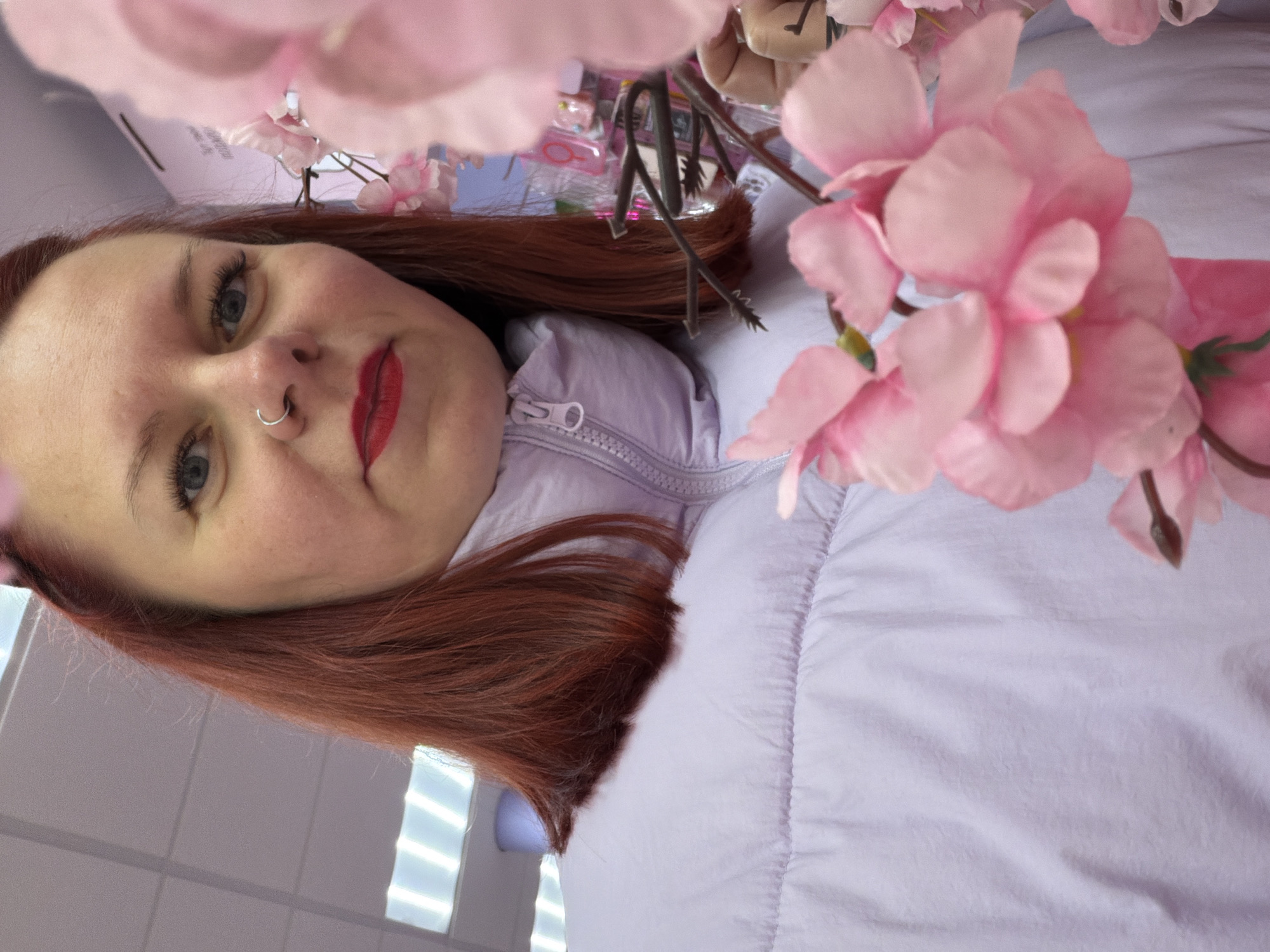 A light-skinned woman with red hair is wearing a light purple down jacket and lipstick while posing with pink flowers in what appears to be an indoor setting in Unknown, Unknown. The scene is brightly lit, and the background is out of focus.