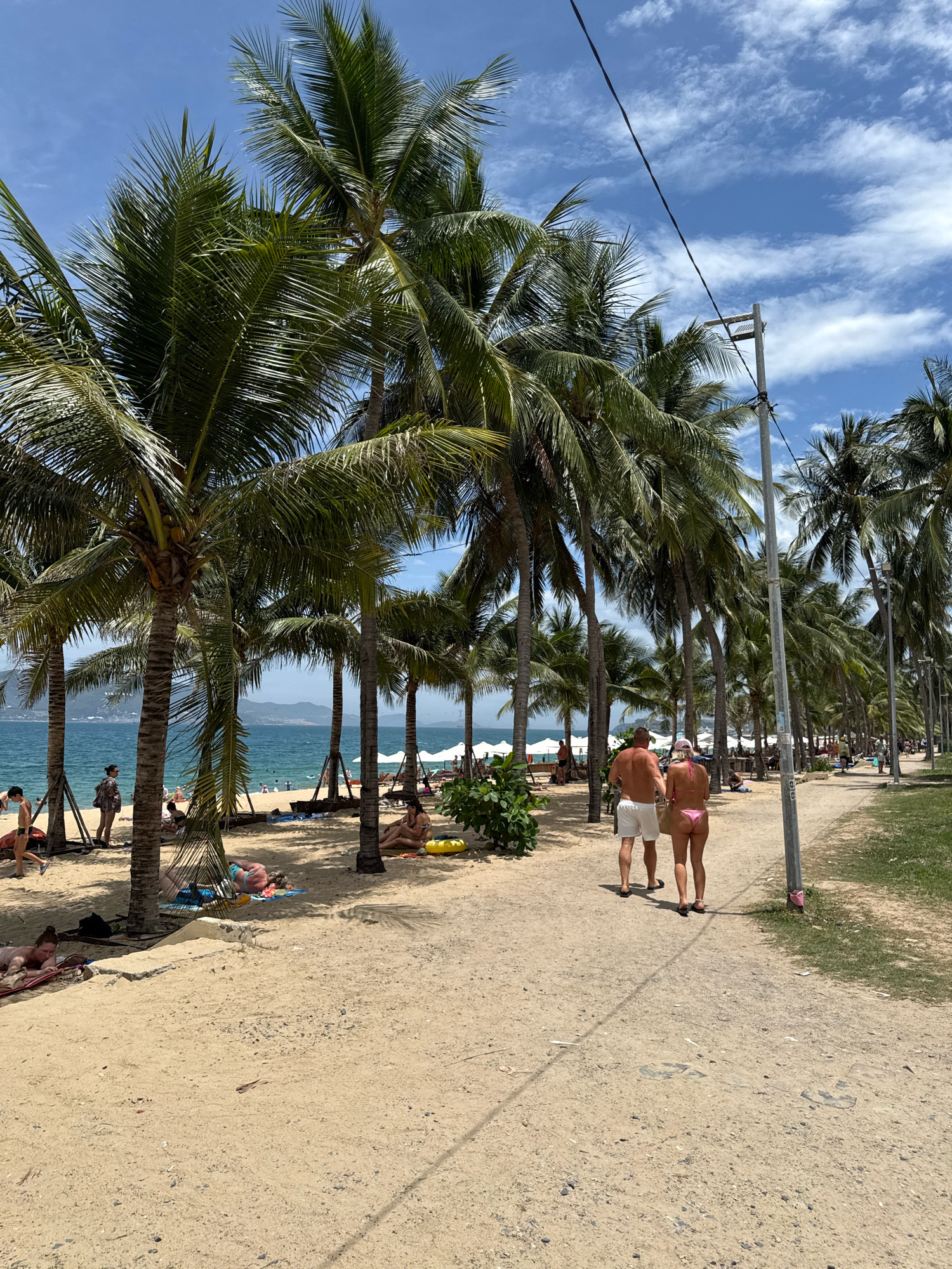 A beach scene in Nha Trang, Vietnam, features a sandy path lined with numerous palm trees. In the foreground, a couple walks away from the camera on the path, with the man wearing white shorts and the woman in a pink bikini. To the left of the path, the beach is populated with several individuals sunbathing on towels and lounge chairs, with some people near the ocean. The ocean itself is visible in the background, with a clear blue sky overhead dotted with white clouds. A utility pole with wires runs along the right side of the path. The overall atmosphere is that of a sunny, tropical vacation destination.