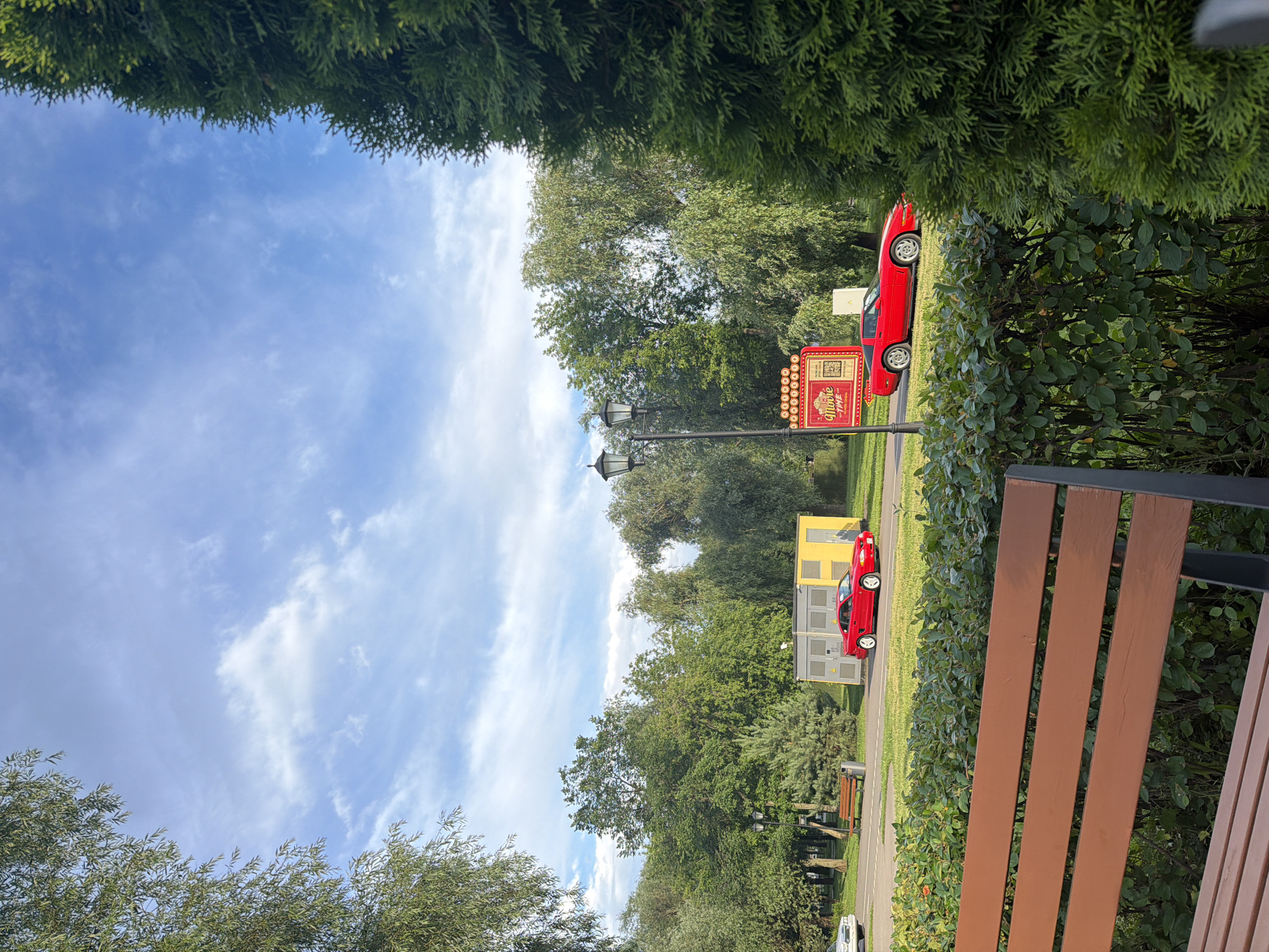 In an outdoor park setting, two red sports cars are parked on a paved surface adjacent to a grassy area and a row of green bushes. A wooden bench is visible in the foreground. In the background, several trees form a dense canopy, with a blue sky and scattered white clouds above. A bright yellow structure and a red sign with "Movie Time" written on it are also present. A lamppost stands near the sign. The scene appears to be a public park or recreation area.